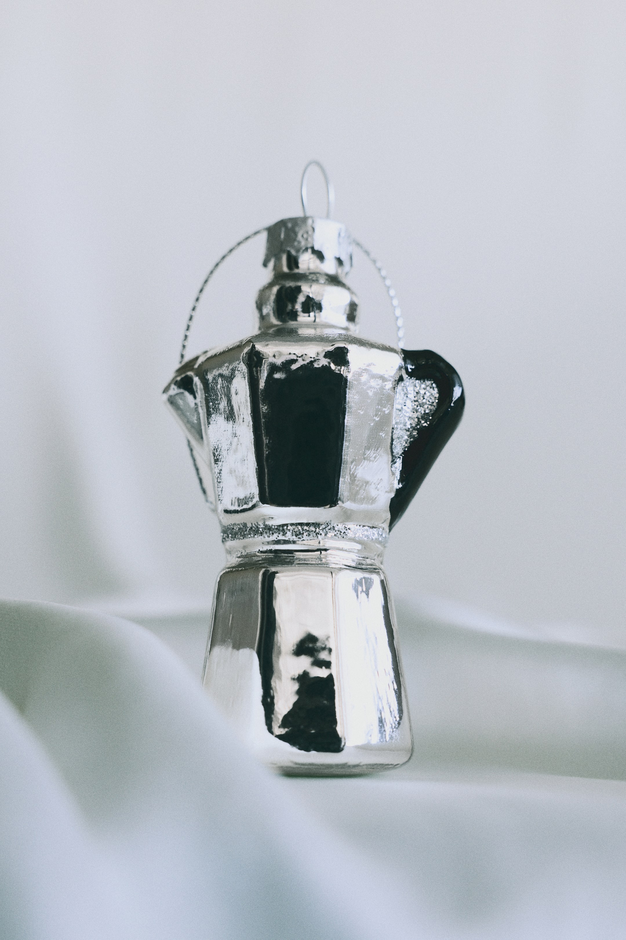 View larger image: A glass Christmas ornament shaped like a moka pot stands upright, featuring a reflective metallic silver finish with a black handle on the side and a silver glitter band around the middle. The ornament is topped with a silver cap and a thin wire hanger loop.