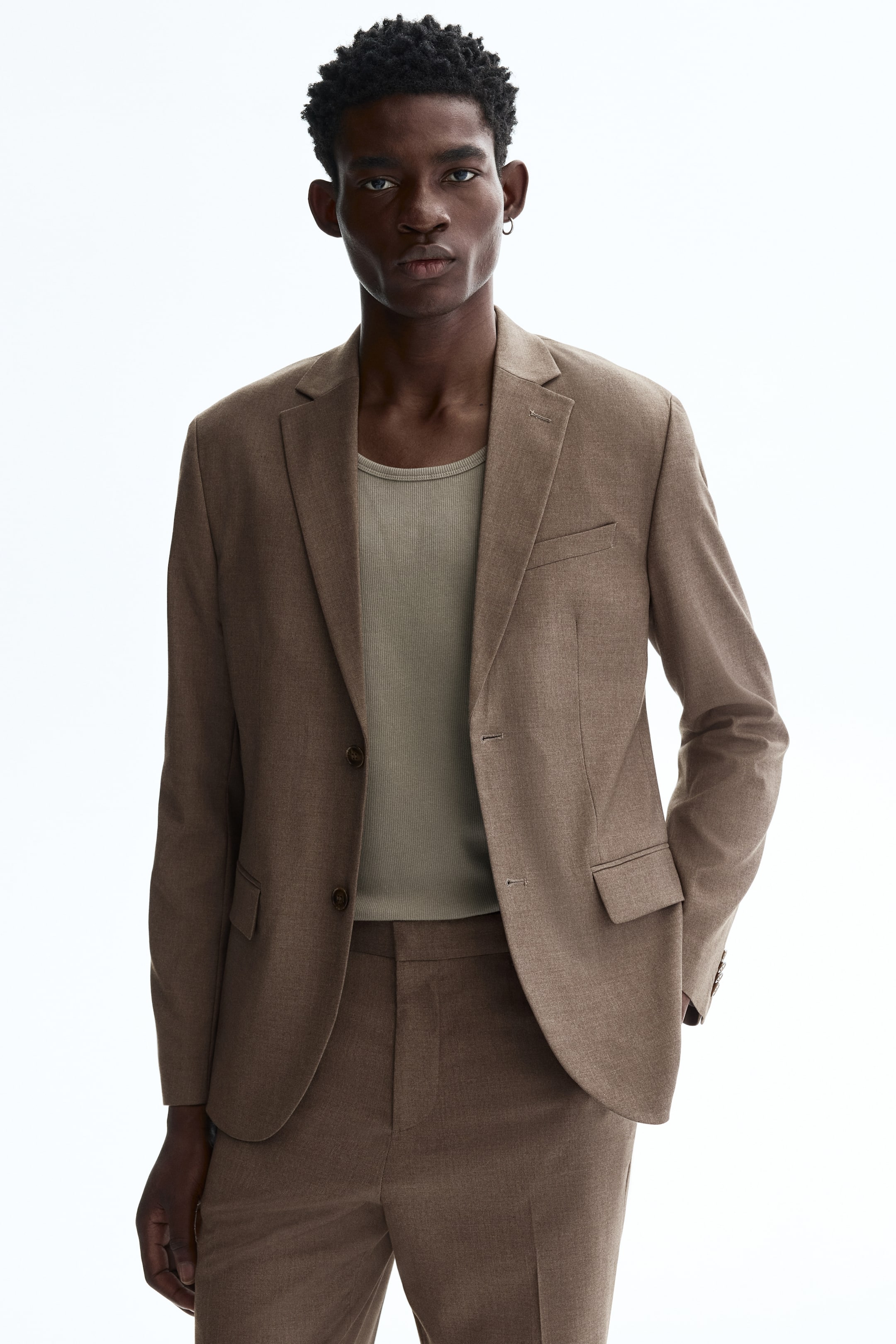 View larger image: The model is wearing a brown single-breasted jacket in a soft weave, featuring notch lapels, a welt chest pocket, and two flap front pockets. It fastens with two front buttons, has decorative cuff buttons, and is layered over a ribbed undershirt.