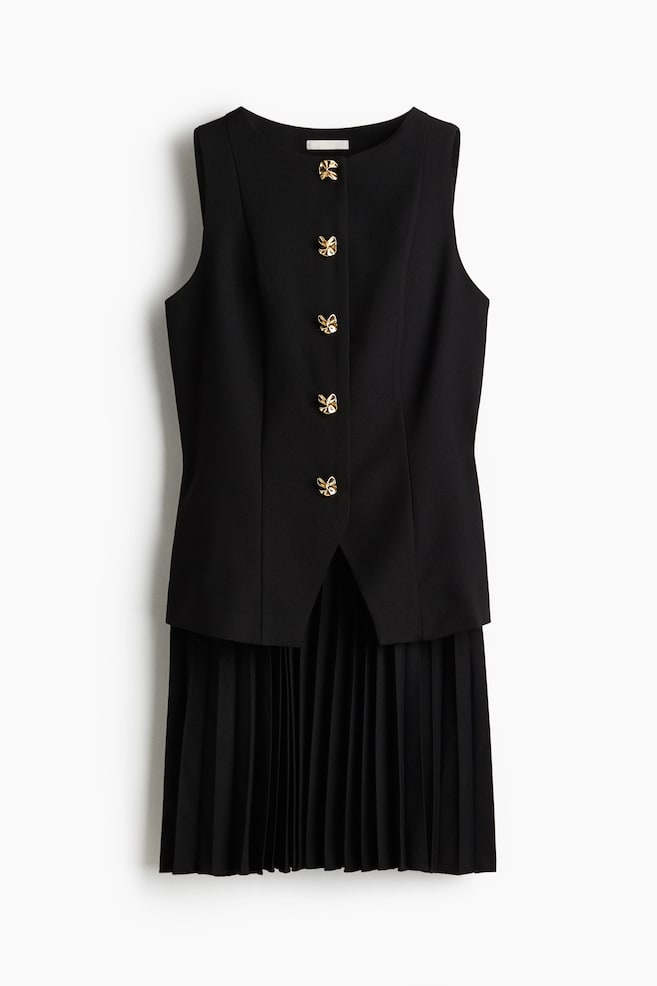 Une robe noire sans manches est présentée de face, avec un col rond et quatre boutons dorés en forme de nœud ornant la patte de boutonnage centrale. Le corsage s'étend en une jupe finement plissée. - 1