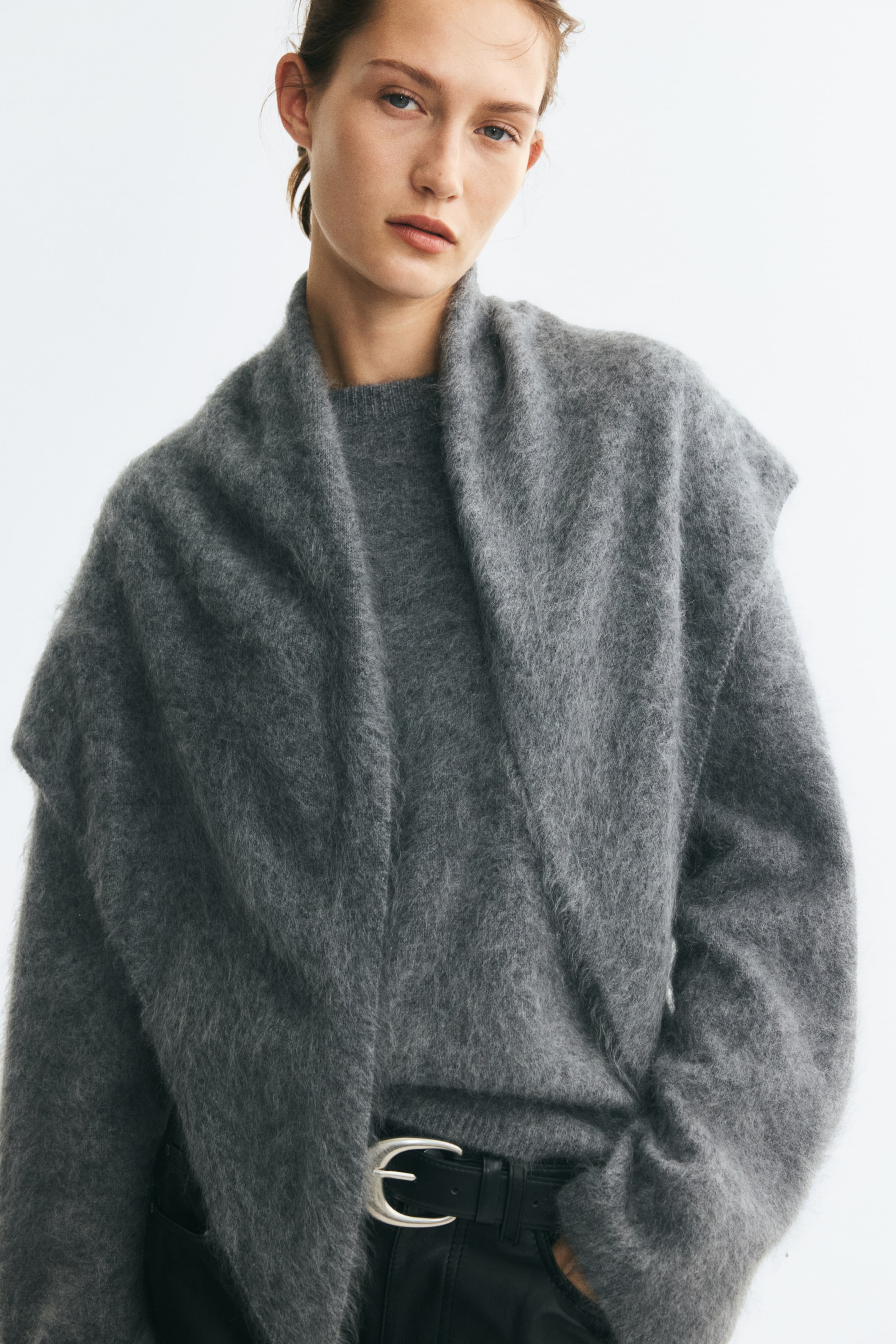 Größeres Bild anzeigen: Das Model trägt einen grauen, gebürsteten Wollstrickpullover und den dazu passenden Dreiecksschal, der locker über die Schultern gelegt ist. Der Pullover verfügt über einen runden, gerippten Ausschnitt und ist in eine schwarze Hose mit einem Gürtel mit silberner Schnalle gesteckt.