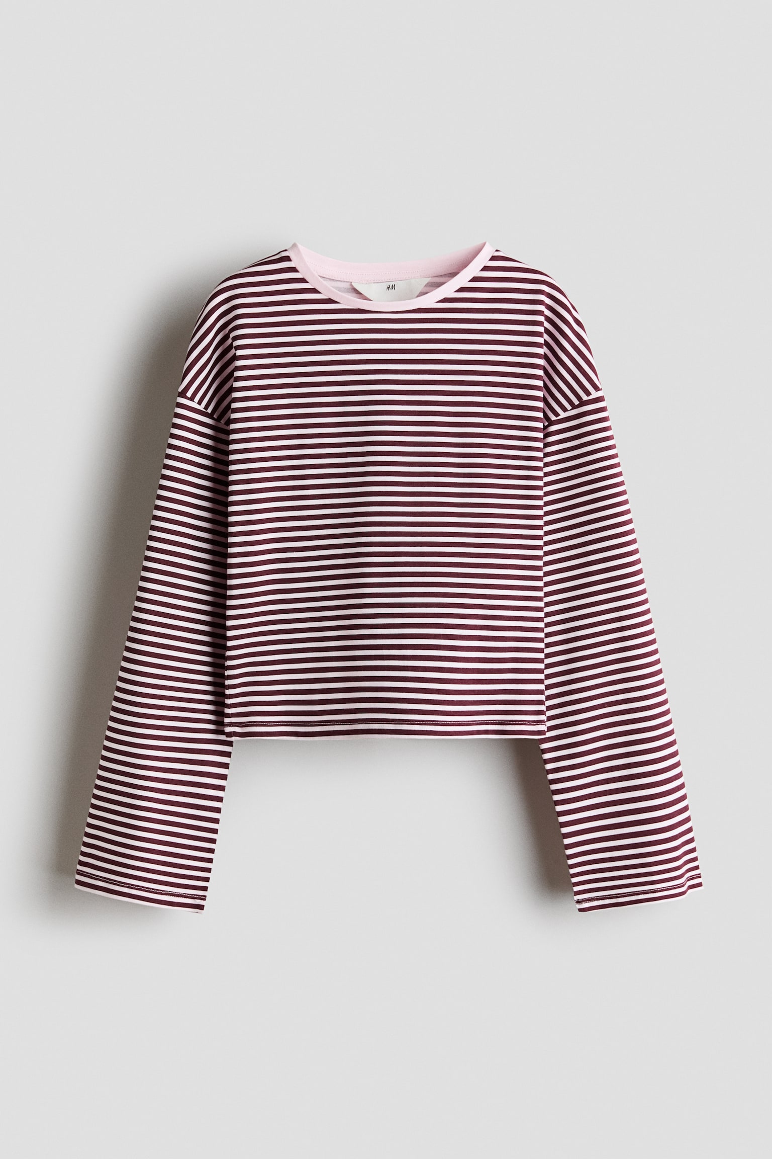 A long-sleeved top is shown from the front, featuring horizontal burgundy and light pink stripes. The top is designed with a solid pink, rib-trimmed round neckline, dropped shoulders, and long, wide sleeves.