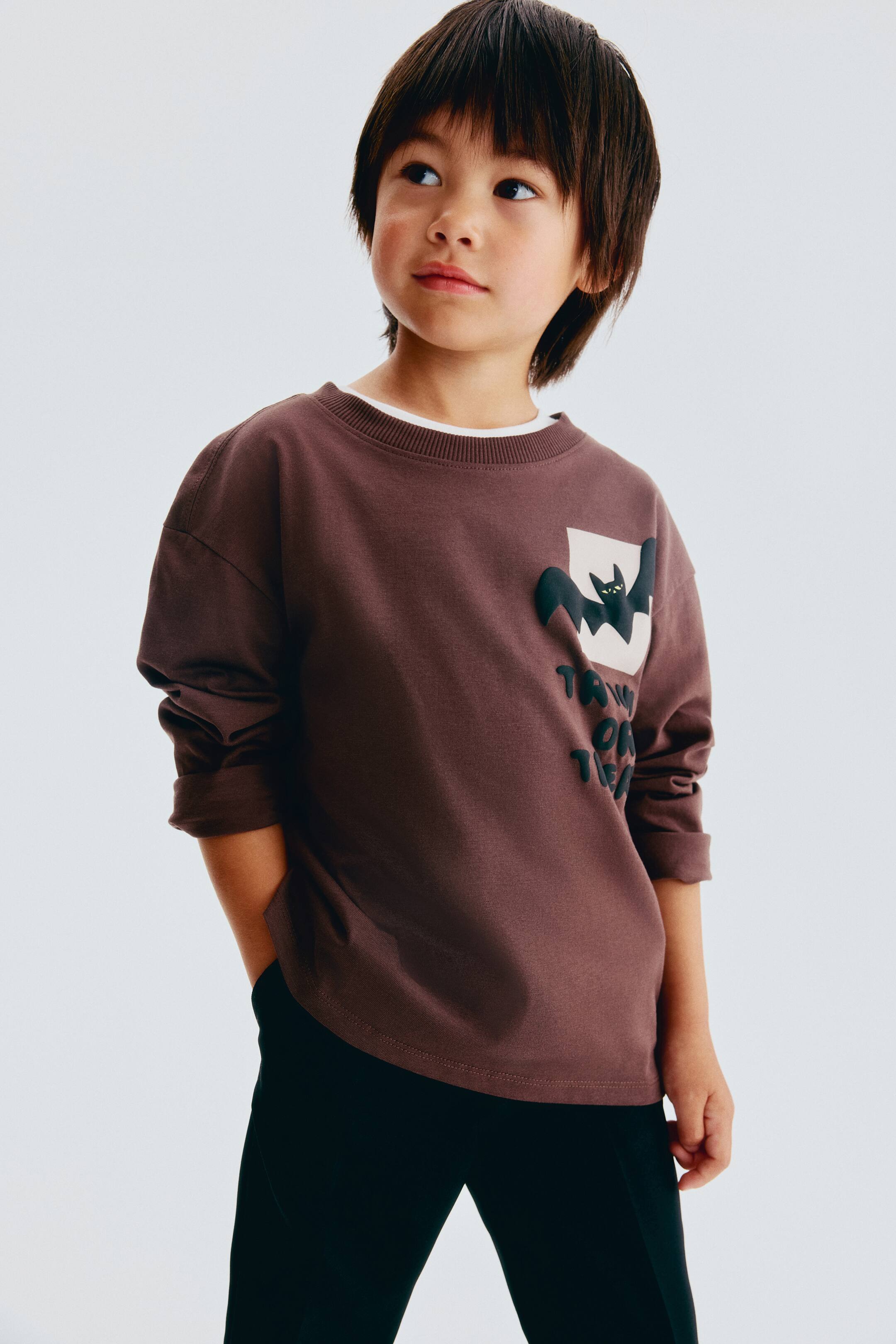 View larger image: A dark brown long-sleeved crewneck shirt is shown from the front, featuring a relaxed fit and a layered white shirt visible at the neckline. The front left chest displays a black bat graphic on a light square, with raised dark lettering partially visible below it.