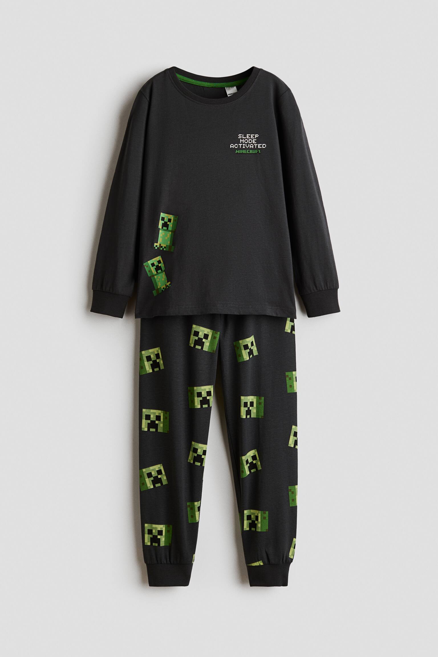 A black two-piece pyjama set is shown from the front, featuring a long-sleeved top and matching trousers. The top features a round ribbed neckline, ribbed cuffs, and green Minecraft graphics, while the trousers feature an all-over print of green pixelated Creeper faces and ribbed cuffs.