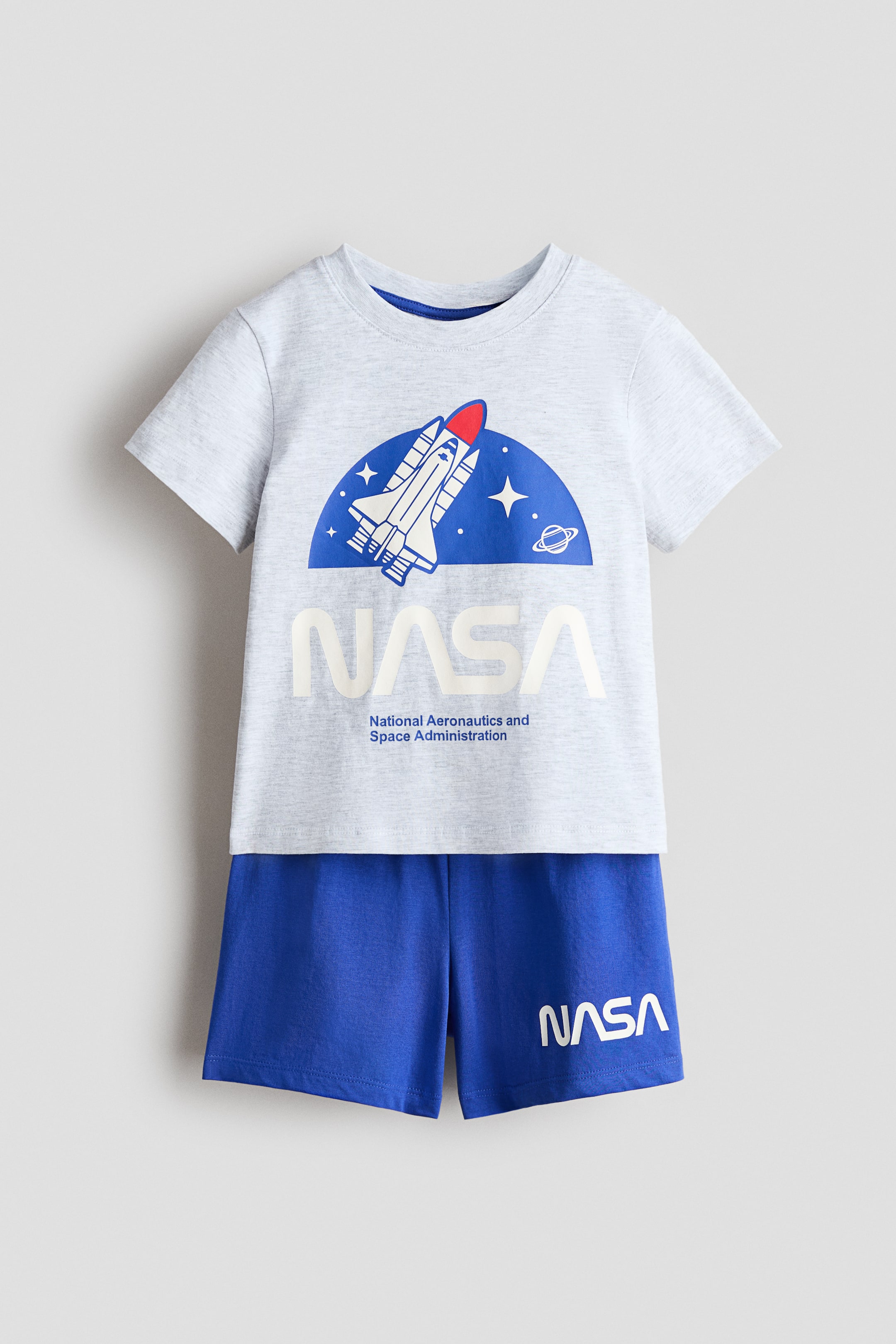 View larger image: A front-facing children's outfit highlights a light grey marl short-sleeved t-shirt paired with royal blue shorts. The t-shirt showcases a blue semi-circle graphic featuring a white and red space shuttle, stars, and a planet, positioned above large white 'NASA' text and smaller 'National Aeronautics and Space Administration' text. The shorts display white 'NASA' text on the left leg.