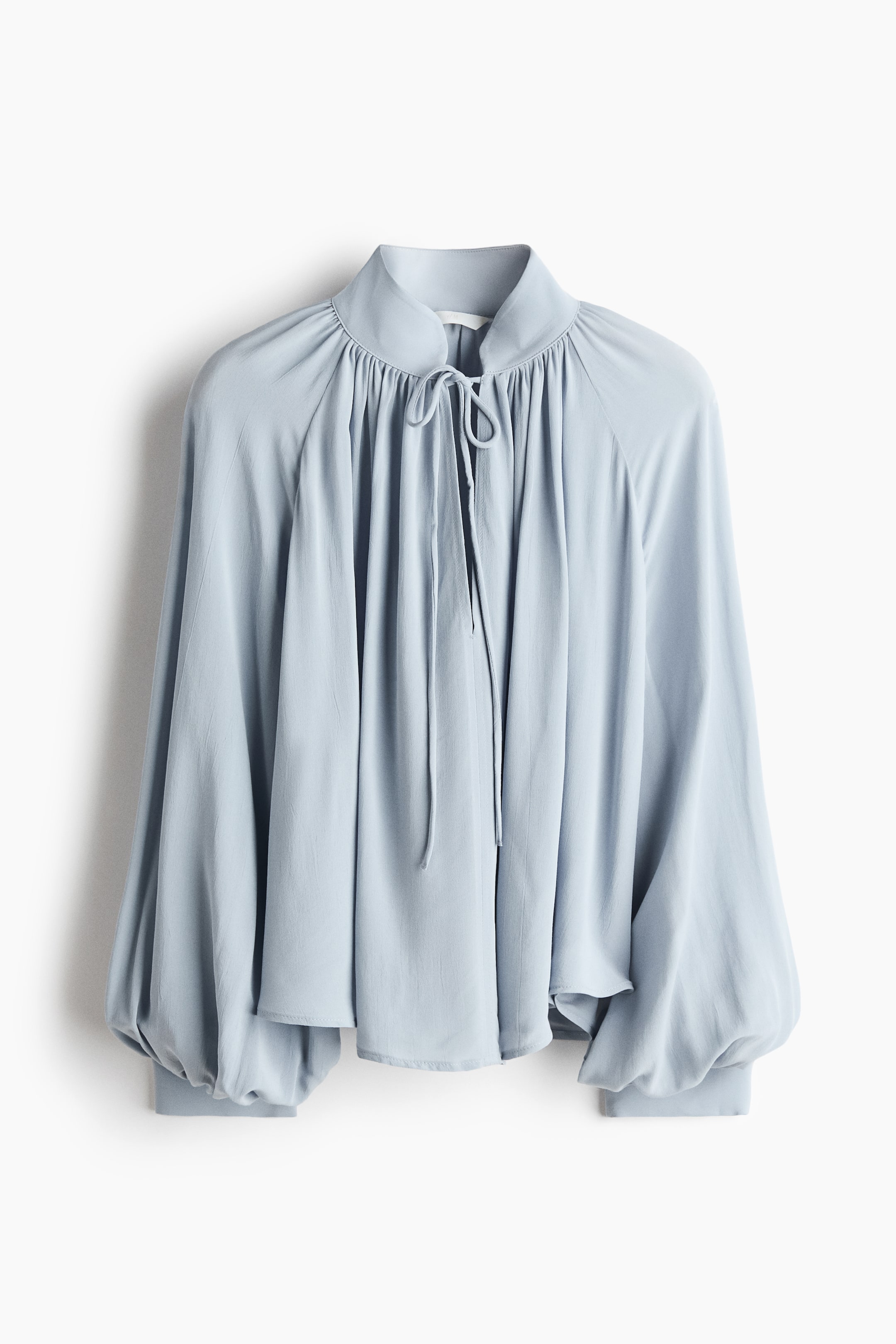 Grotere afbeelding bekijken: Een lichtblauwe blouse met een hoge, geplooide halslijn en een sluiting met trekkoord aan de voorkant. De blouse heeft lange raglanmouwen die bij de manchetten zijn geplooid, wat zorgt voor een ontspannen, volumineus silhouet.