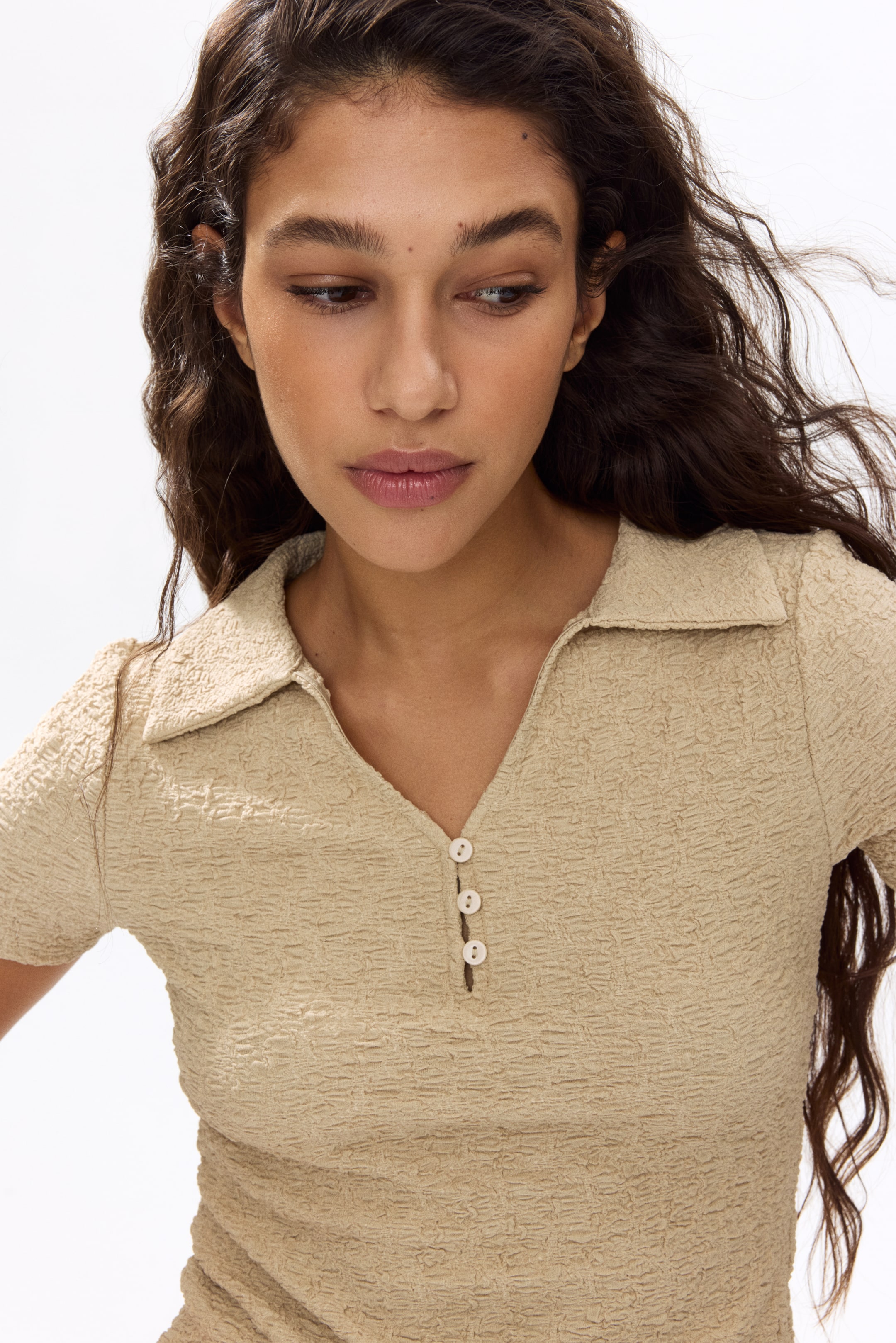 View larger image: A beige crinkle-textured short-sleeved top featuring a pointed collar, a V-neck opening with a three-button placket, and slightly puffed sleeves.