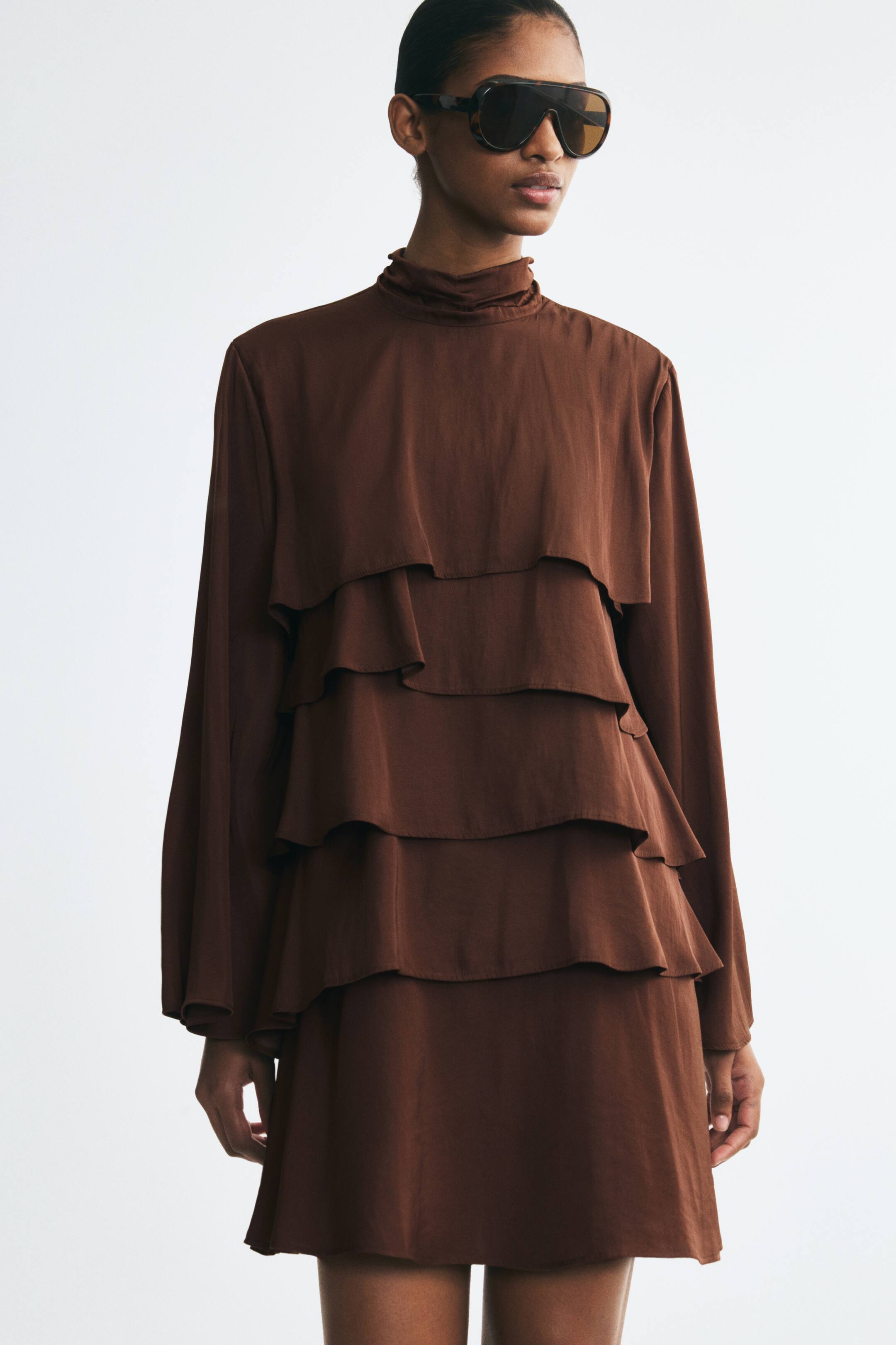 View larger image: A short, brown, loose-fit dress in softly draping fabric is being worn by the model, featuring a high ruched stand collar, long wide sleeves, and multiple flounced tiers cascading down the front. The dress is styled with oversized, dark sunglasses.