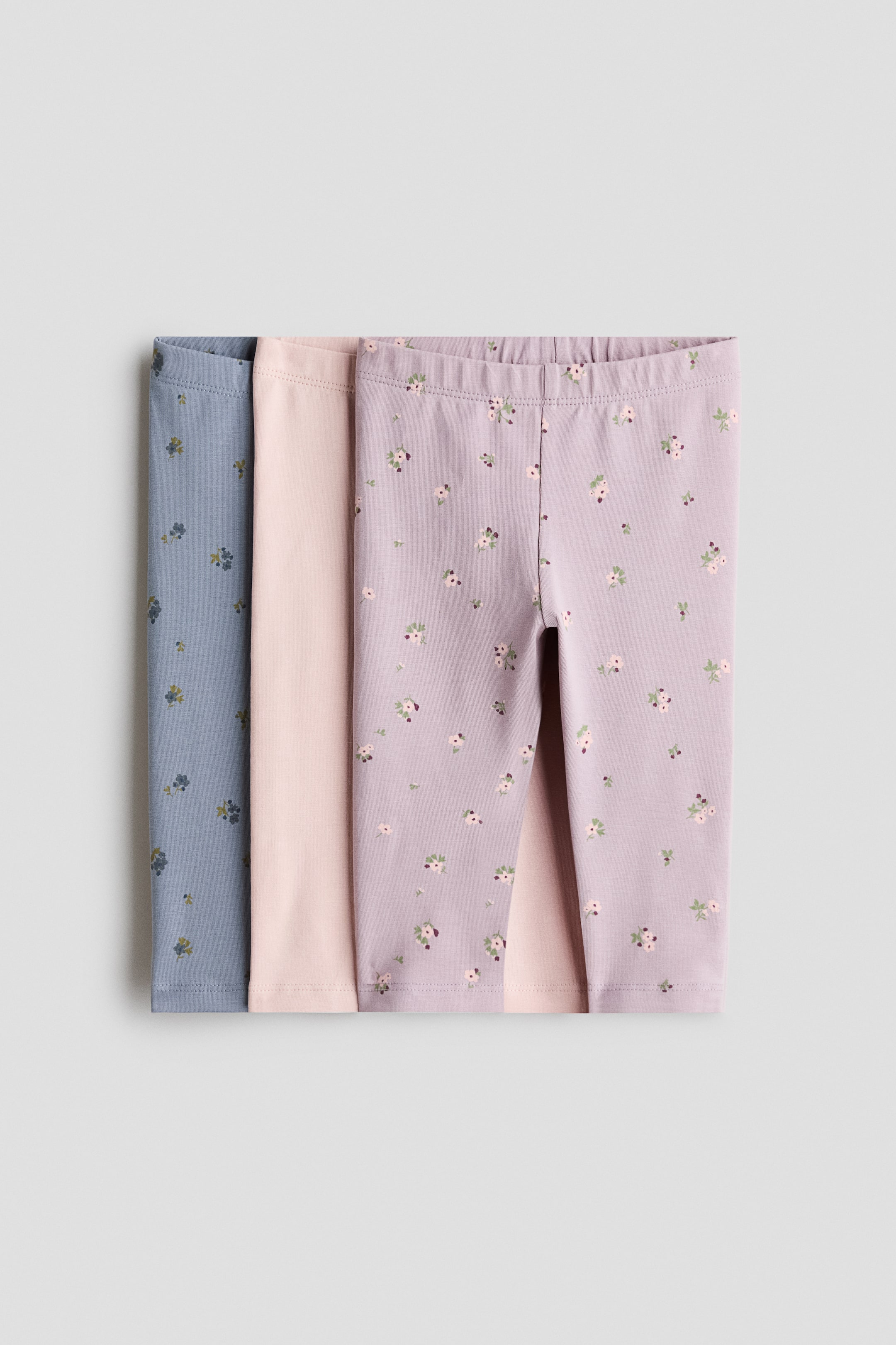 View larger image: Three pairs of soft cotton jersey leggings are shown from the front. The top pair is purple with a small floral print and an elasticated waist. Behind it, a plain pink pair and a blue pair with a matching floral print are layered, also with elasticated waists.