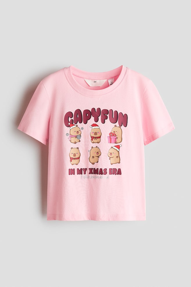 A light pink cotton jersey t-shirt, facing front, is featuring a ribbed round neckline and short sleeves. The front is displaying a graphic design with the word "CAPYFUN" above six cartoon capybaras in festive accessories and the phrase "IN MY XMAS ERA" printed underneath. - 1