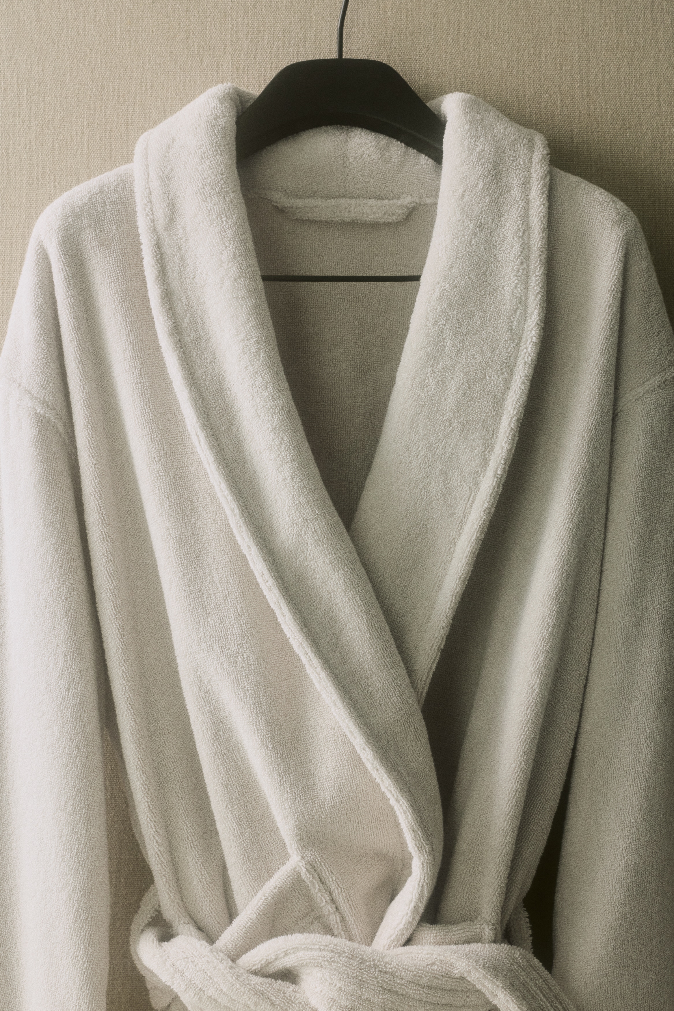 A light grey terry dressing gown is shown from the front, featuring a shawl collar, long sleeves, and two large patch pockets. A matching terry tie belt cinches the waist and is tied in a knot, while woven trim details both the cuffs and hem.