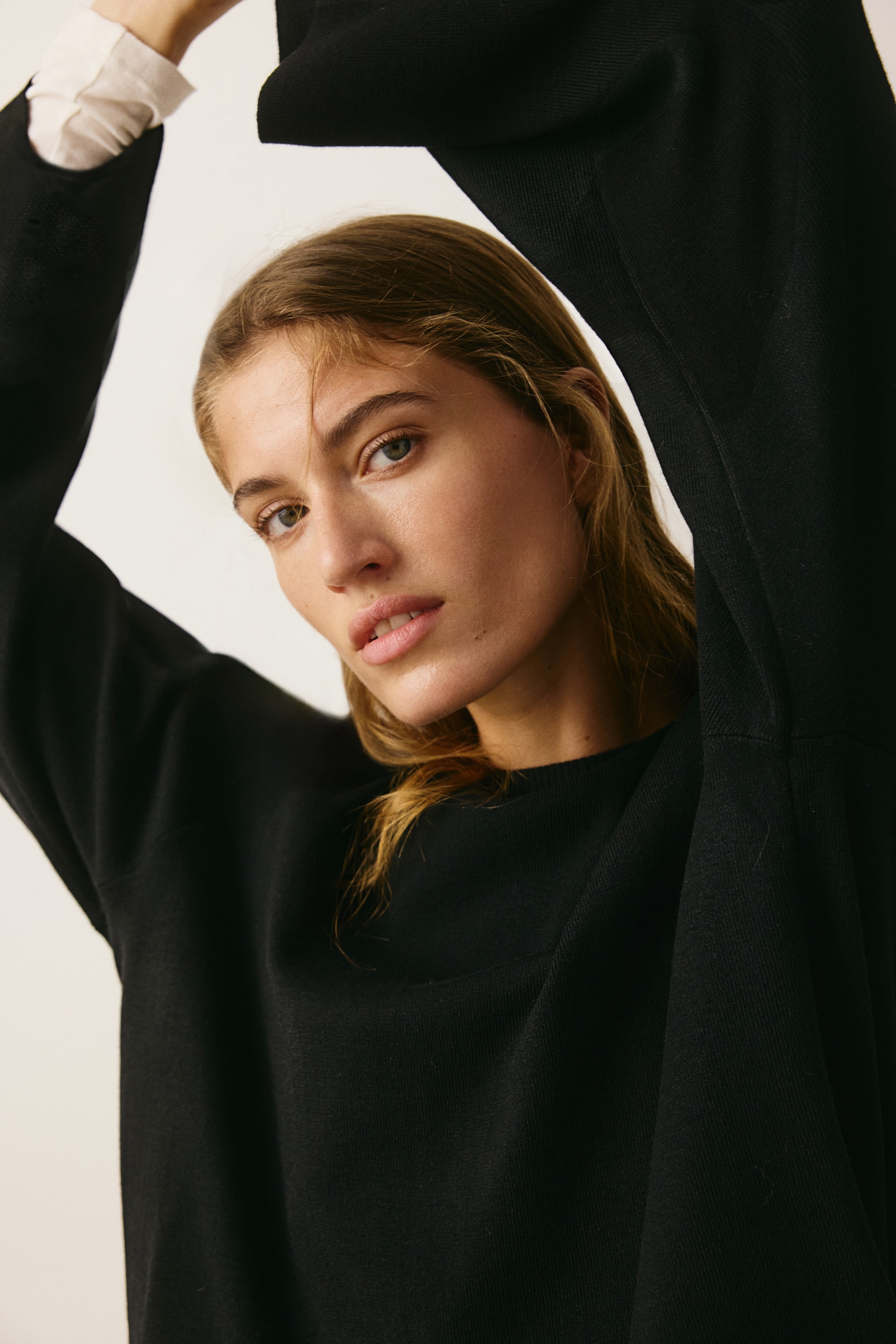 View larger image: A black, fine-knit oversized sweater is featured from the front, highlighting its ribbed crew neck and dropped shoulders. The loose-fitting long sleeves drape as they are raised, revealing a light-colored underlayer cuff.