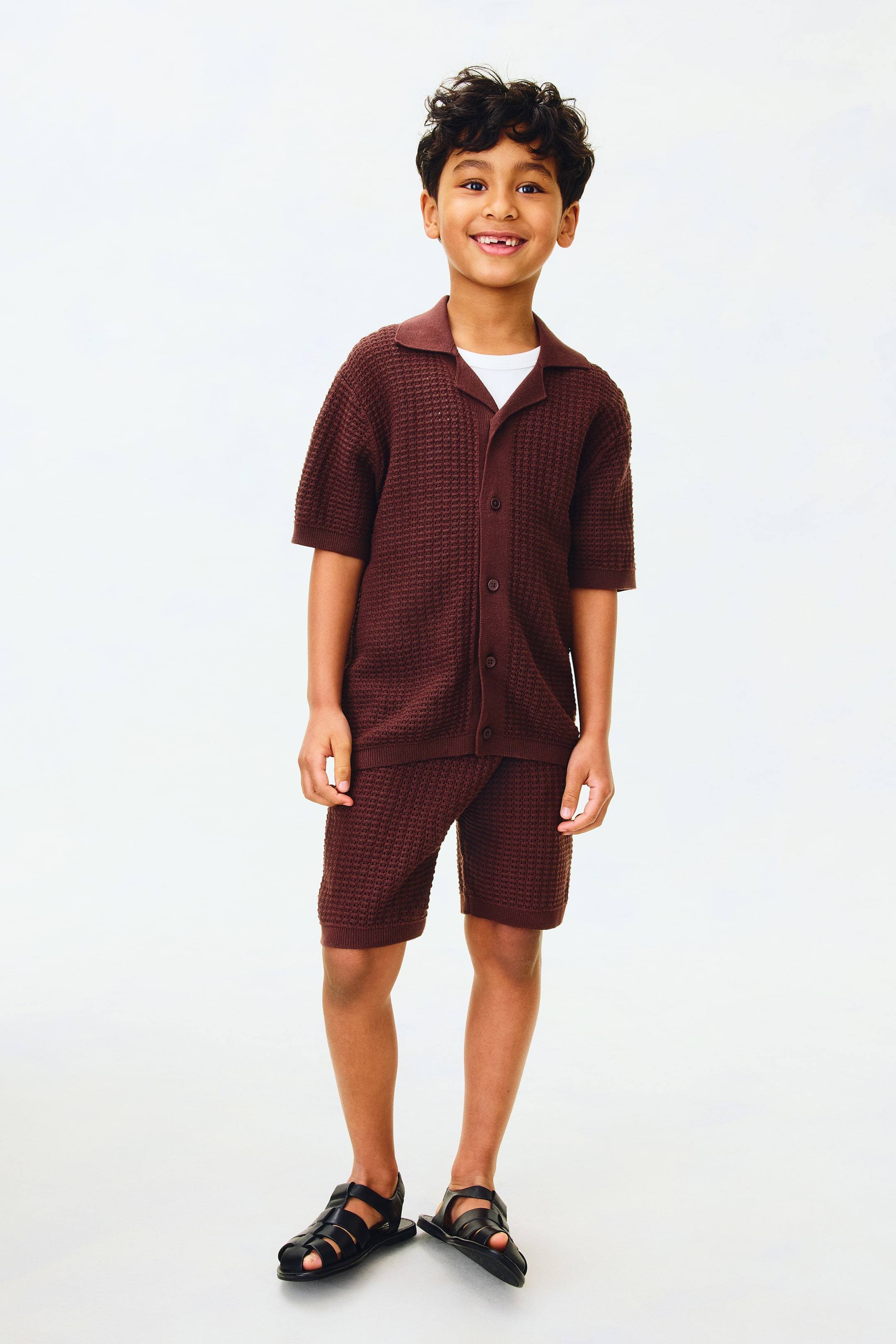 Größeres Bild ansehen: Ein dunkelbraunes, kurzärmeliges Polohemd aus strukturiertem Strick mit Knopfleiste wird über einem weißen T-Shirt mit Rundhalsausschnitt getragen, kombiniert mit passenden Strick-Bermudashorts und schwarzen Riemchensandalen.