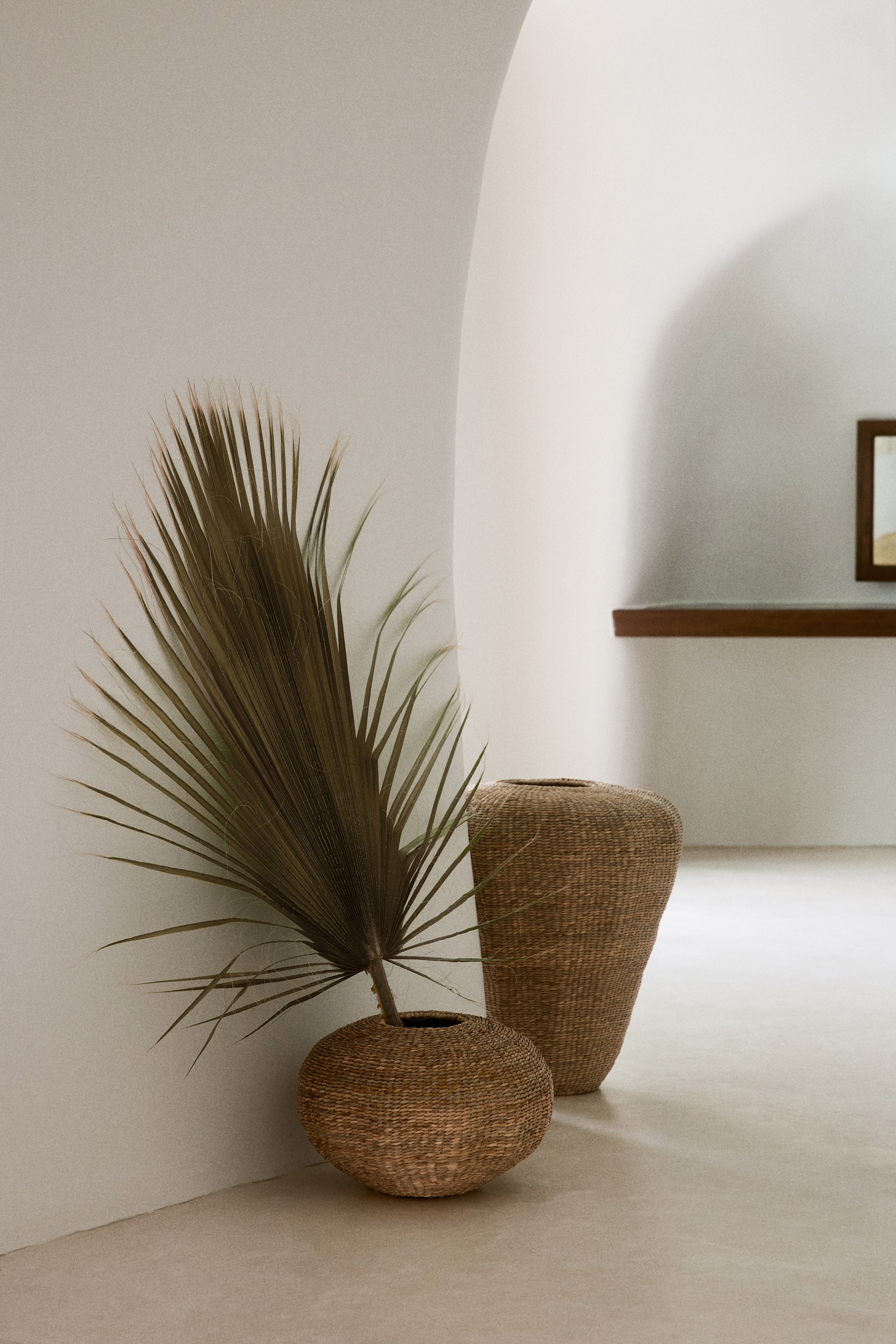 View larger image: Two woven baskets are placed on the floor. A large, dried fan palm frond is positioned in the smaller, rounded woven basket, while a taller, tapered woven basket stands beside it.