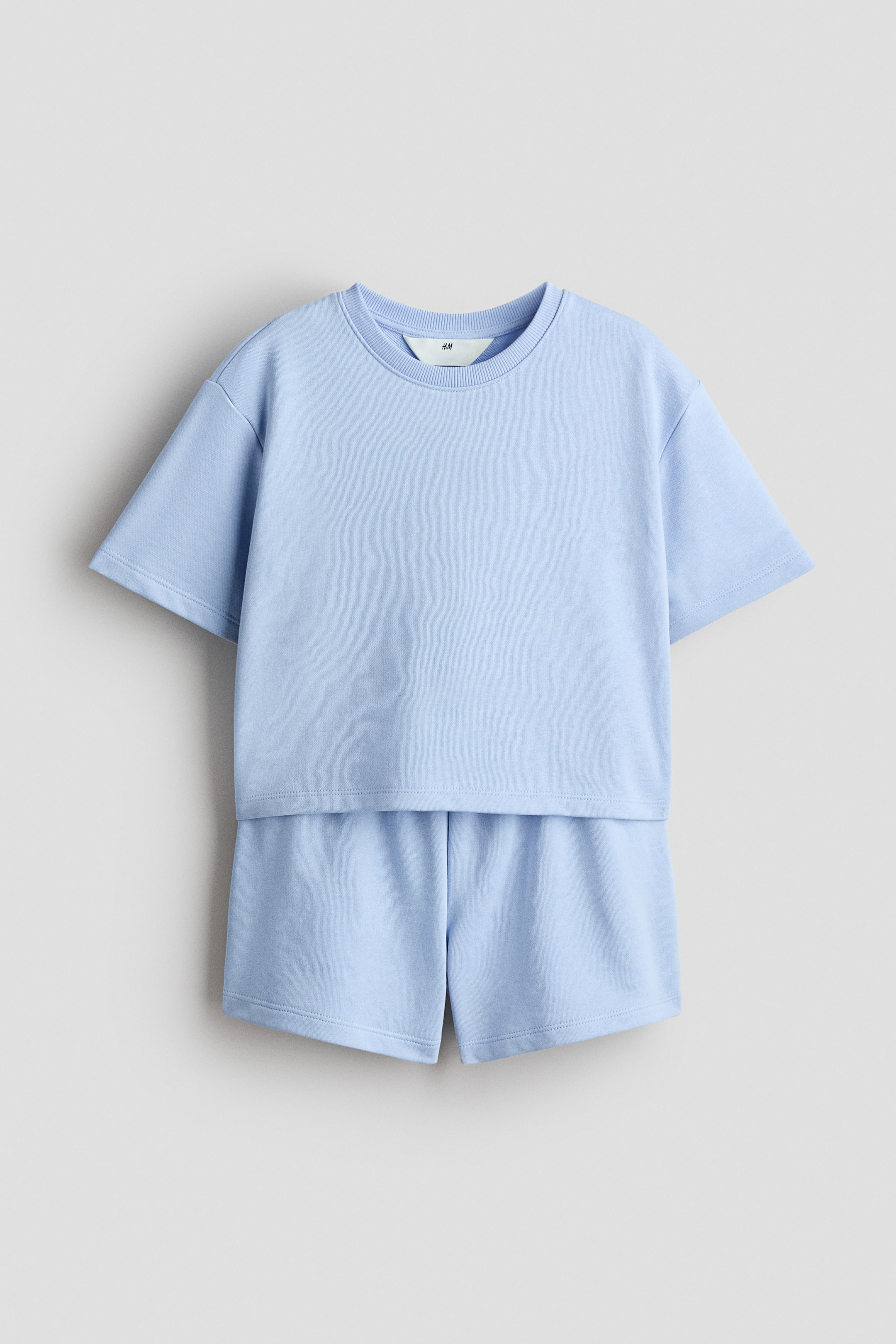 Größeres Bild anzeigen: Ein hellblaues, bequemes Set aus leichtem Sweatshirtstoff, bestehend aus einem T-Shirt und Shorts, wird von vorne präsentiert. Das T-Shirt ist mit einem runden, gerippten Ausschnitt, überschnittenen Schultern und einem geraden Saum mit kurzen Schlitzen an jeder Seite designt. Die Shorts zeigen eine verdeckte Elastik am Bund.