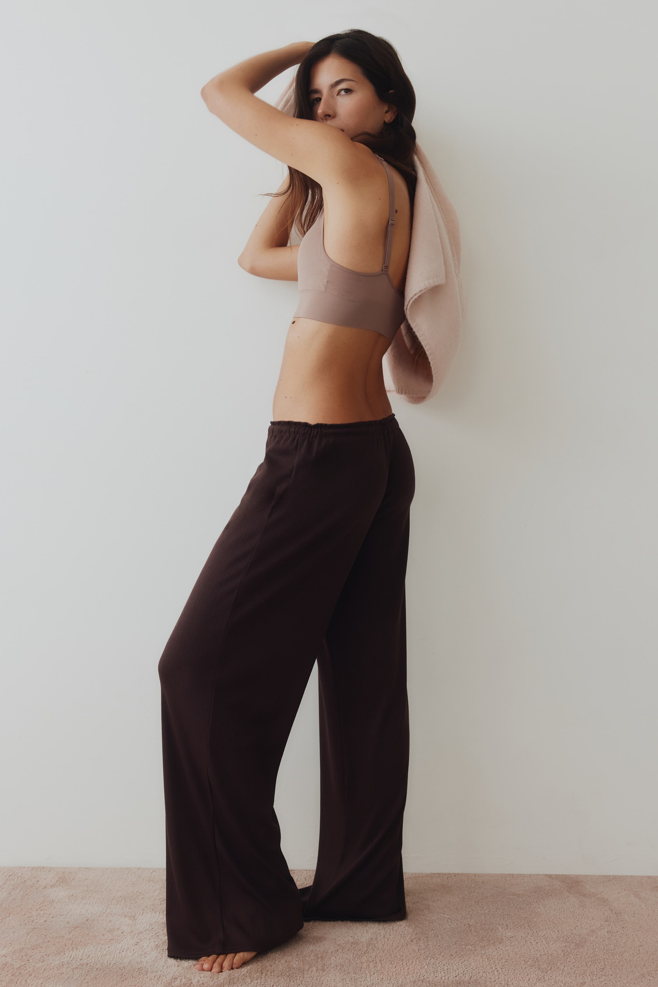 View larger image: Dark brown wide-leg pants with a relaxed fit and an elastic waistband, shown from a side view. A light brown bralette with thin, adjustable shoulder straps and a smooth underband, visible from a side-back angle.
