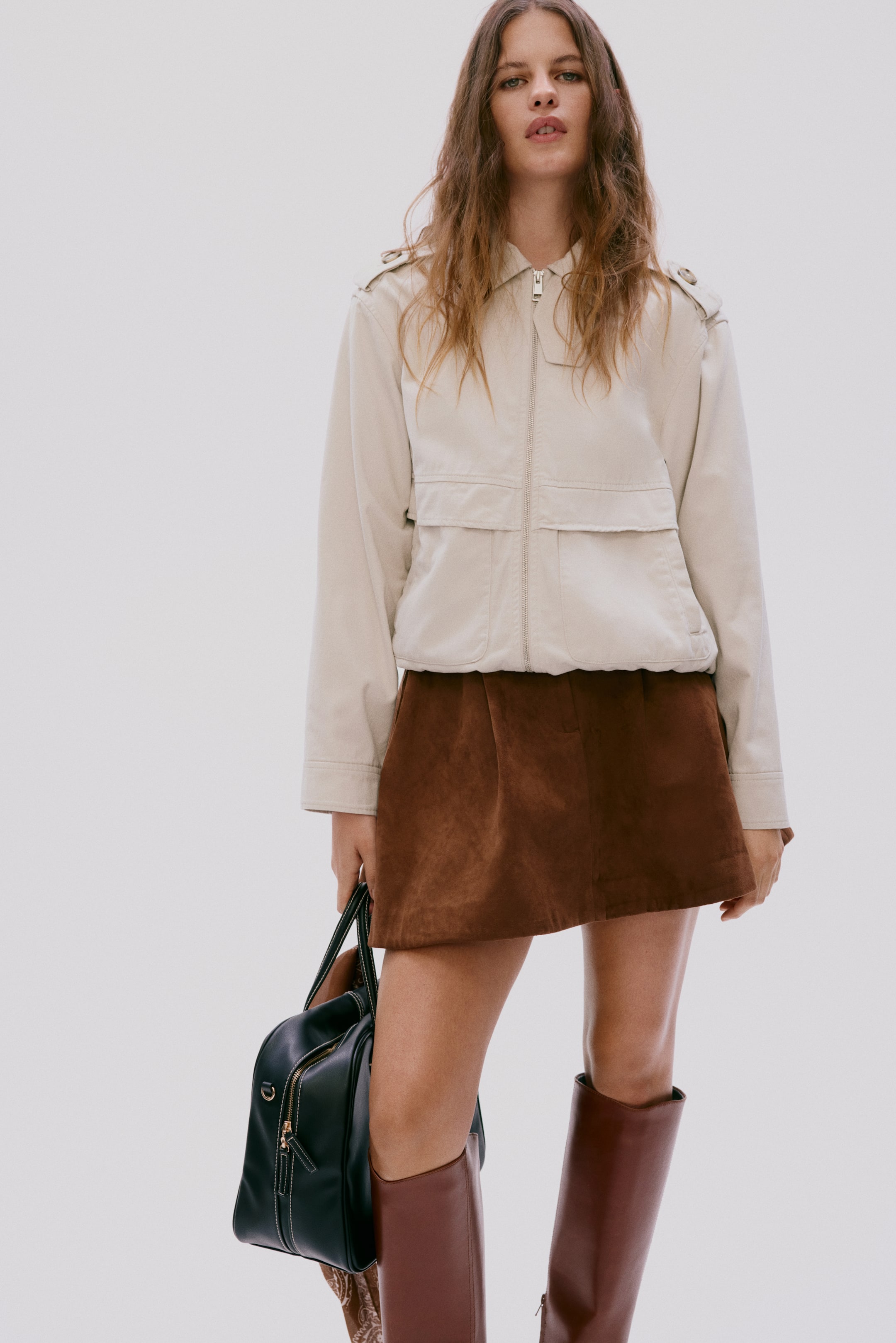 הצגת תמונה גדולה יותר: ז'קט בומבר בצבע בז' בהיר כולל רוכסן קדמי מלא, צווארון עומד ולשוניות כתפיים. שני כיסי דש נראים על החזה, יחד עם שני כיסים טלאים תחתונים ושוליים אלסטיים. נלבש מעל חצאית מיני דמוית זמש חום.