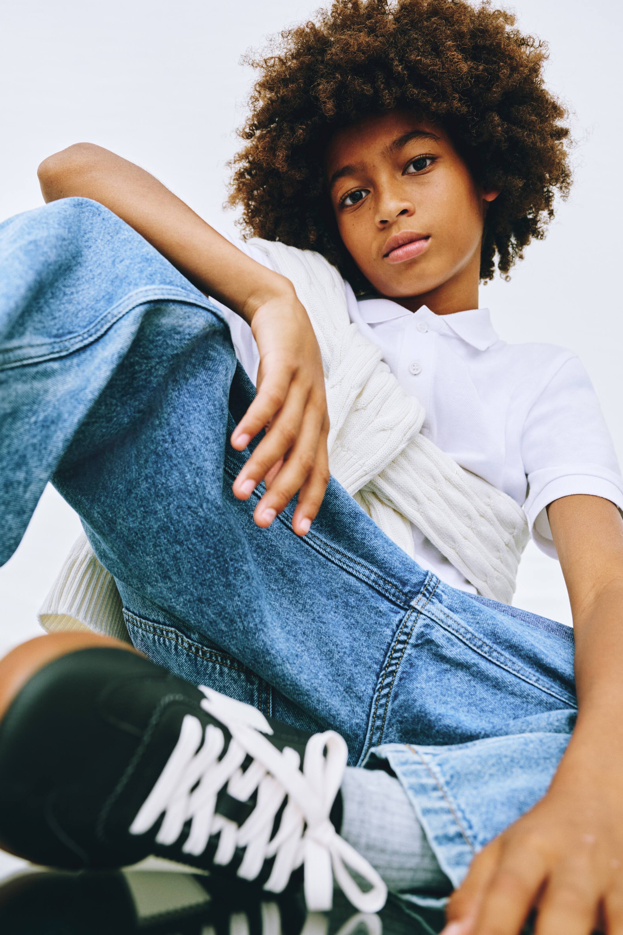 Grotere afbeelding bekijken: Relaxte jeans in een middelblauwe wassing worden gedragen in een frontaal, laag standpunt, gestyled met een wit polohemd met korte mouwen en een witte kabelgebreide trui die over de schouders is gedrapeerd. De jeans hebben klassieke contrasterende stiksels en een wide-leg silhouet.