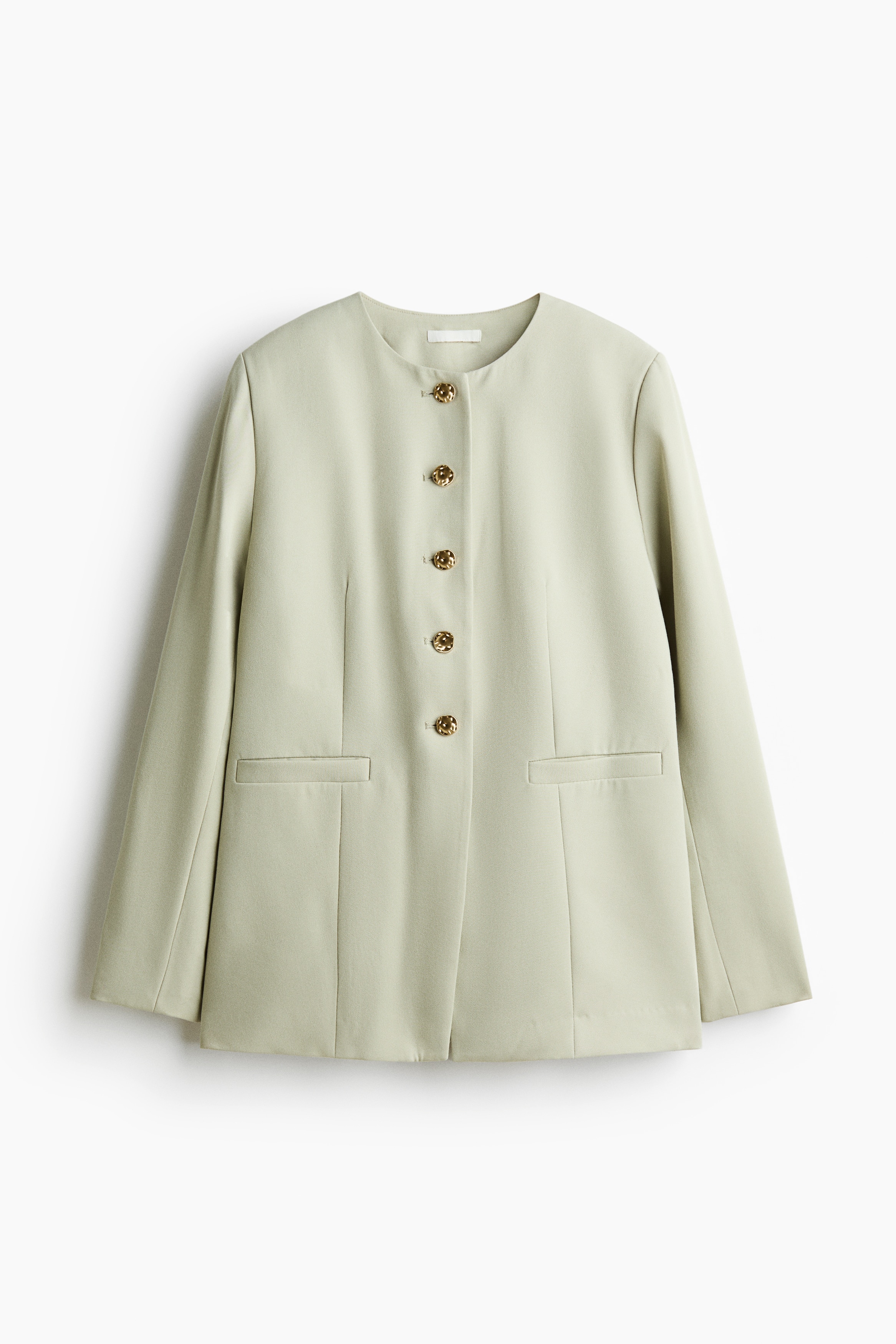 The model is wearing a long, single-breasted twill blazer in a light sage green, featuring a collarless round neckline, structured shoulders, and irregular-shaped gold metal buttons along the front. The blazer is styled with matching trousers.