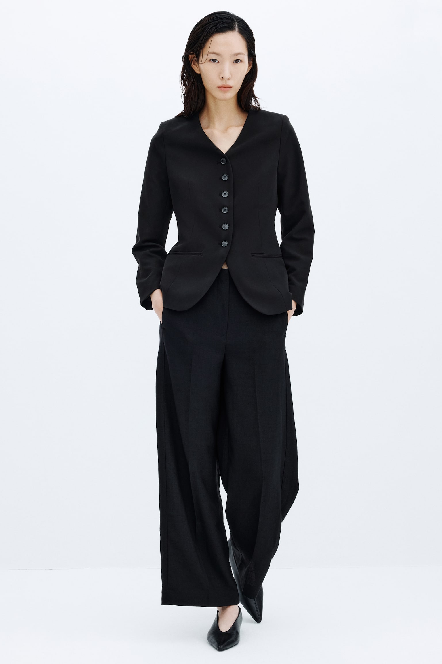 A black relaxed-fit blazer is hanging facing front, featuring a lapelless, deep V-neckline and a front closure with six black buttons. The blazer shows vertical waist darts, long sleeves, and two horizontal jetted pockets at the hips. - 1