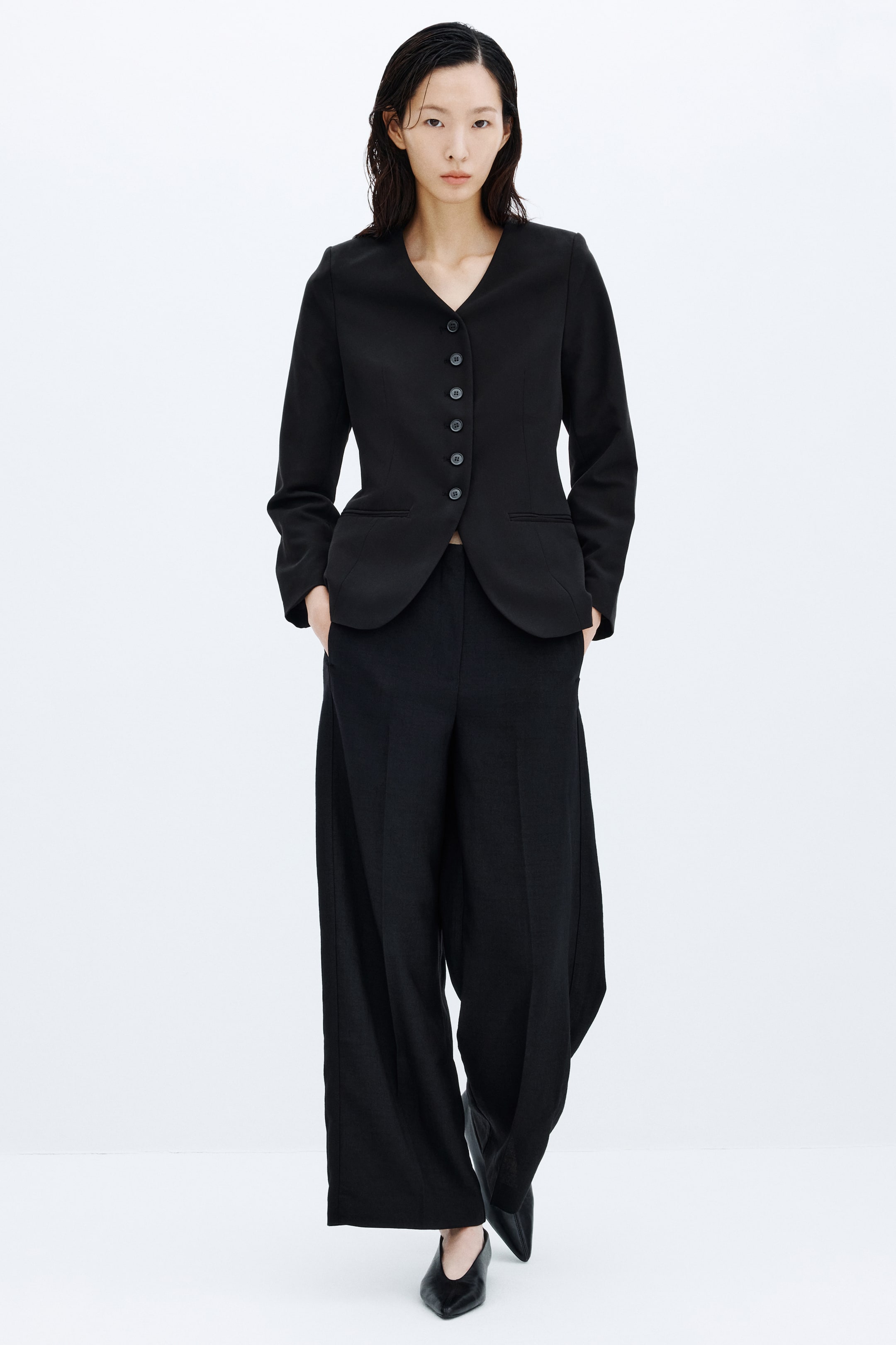 View larger image: A black lapelless twill blazer featuring a V-neckline, front button closure, waist darts, and jetted pockets is worn by the model. The blazer is styled with matching wide-leg black trousers and black pointed shoes.