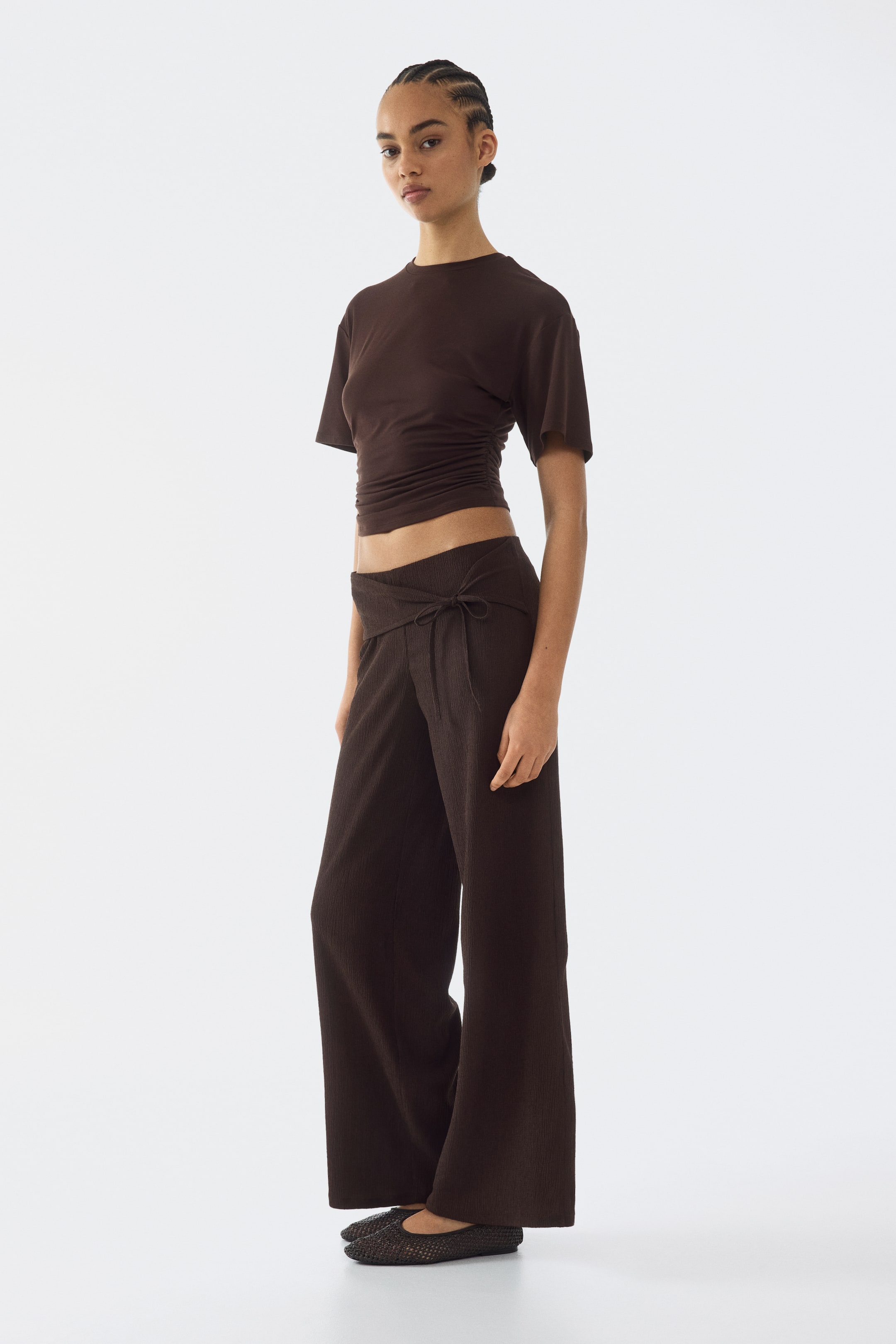 Ver imagem maior: Um top castanho-escuro de manga curta com laterais franzidas e calças largas a condizer estão a ser apresentadas, ligeiramente anguladas para a direita do espectador. As calças apresentam uma cintura com laço envolvente e uma textura subtil canelada.
