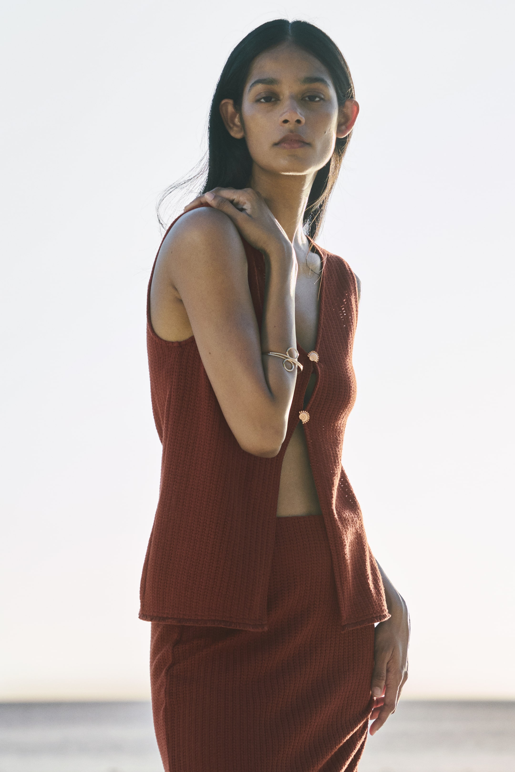 View larger image: "A rust-coloured knitted two-piece set showcases a sleeveless vest adorned with decorative spiral buttons along the front, worn partially open, paired with a matching knitted skirt."