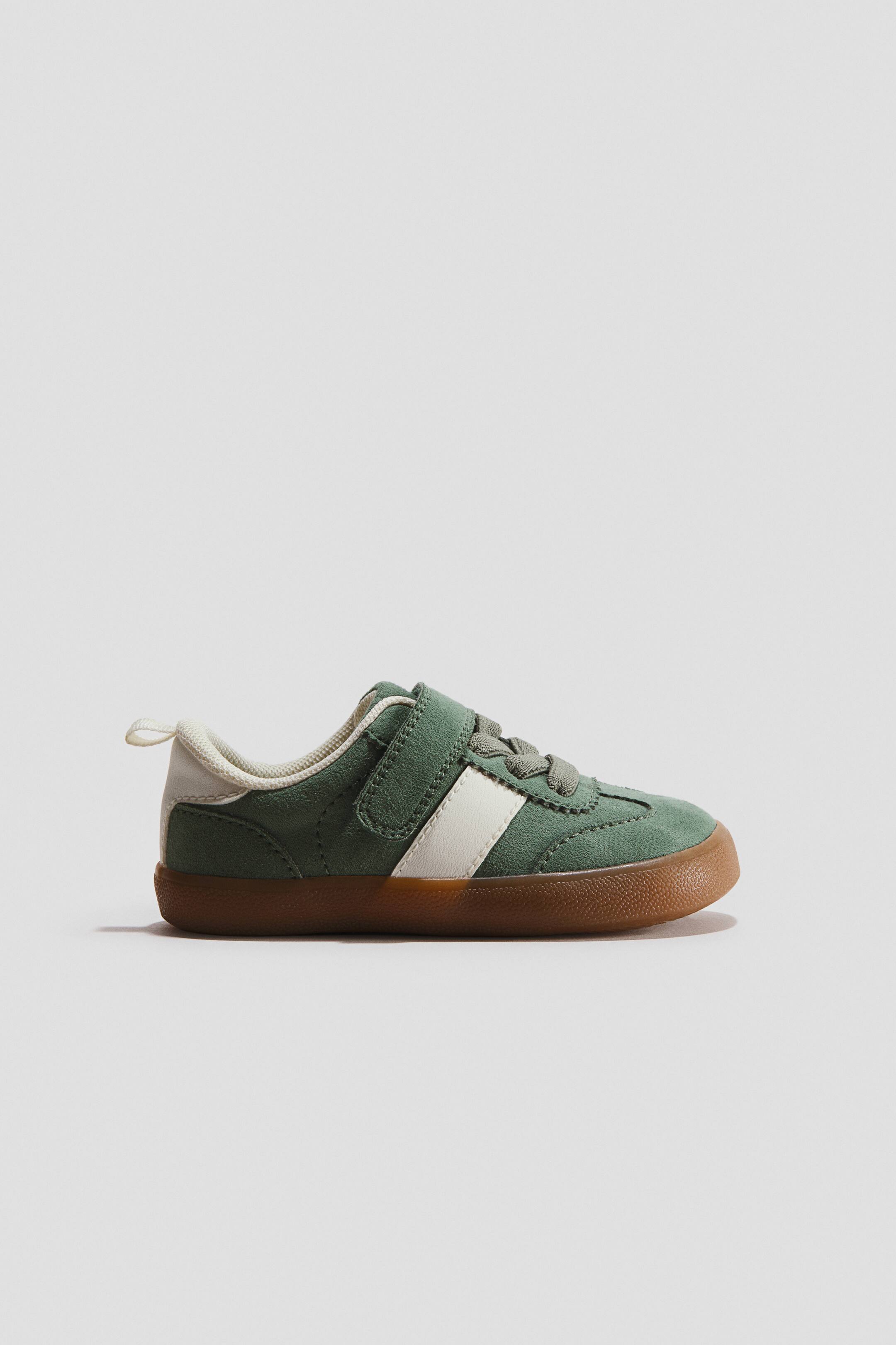 View larger image: Barefoot trainers are shown in a side profile facing right, featuring a green suede upper with a diagonal cream stripe and a hook-and-loop strap closure. The shoe features a thin, flexible gum sole that wraps slightly around the toe and heel, finished with a cream-coloured lining and heel tab.