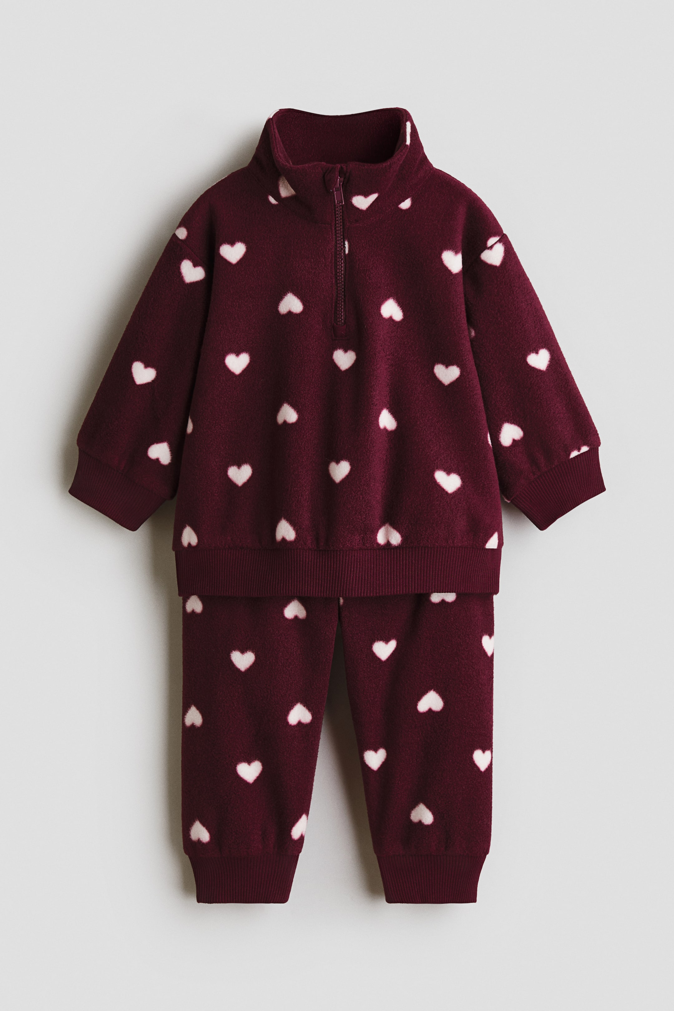View larger image: A burgundy fleece two-piece set features white heart patterns throughout. The front-facing top shows a quarter-zip closure, a stand-up collar, ribbed cuffs, and a ribbed hem, while the matching pants display full-length legs with ribbed ankle cuffs.