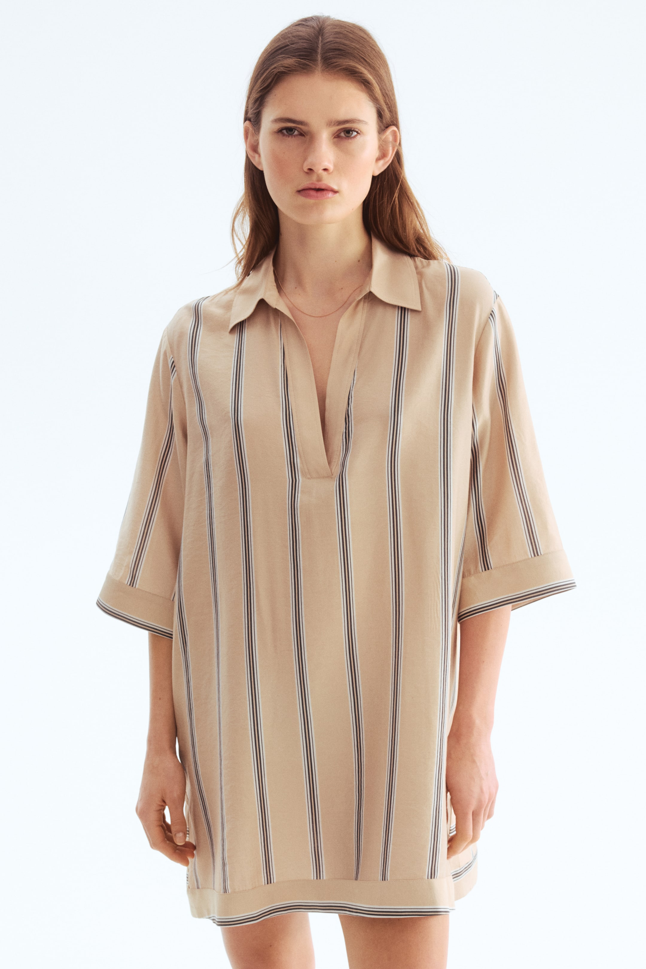 View larger image: A beige tunic dress adorned with vertical stripes, each stripe showcasing a wider dark line paired with a thinner light line, presents a relaxed, straight silhouette when viewed from the front. Crafted with a pointed collar, a V-neck placket, and three-quarter length wide sleeves featuring folded cuffs, this dress is both stylish and comfortable.
