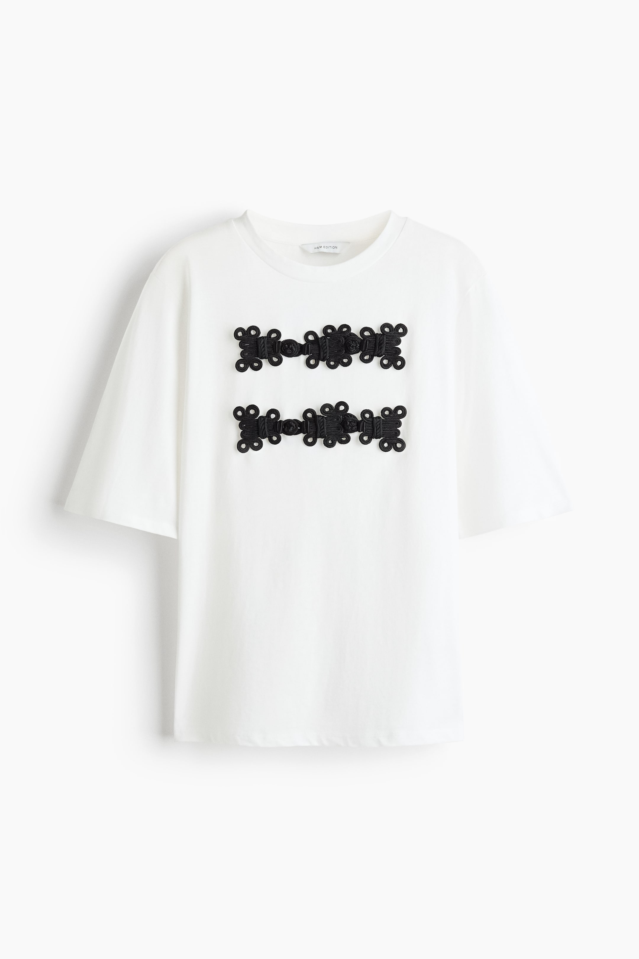 Zobacz większy obraz: Biały T-shirt z okrągłym dekoltem jest ustawiony przodem, z luźnymi krótkimi rękawami i dwiema poziomymi rzędami dekoracyjnych czarnych, plecionych zapięć typu frog na piersi.