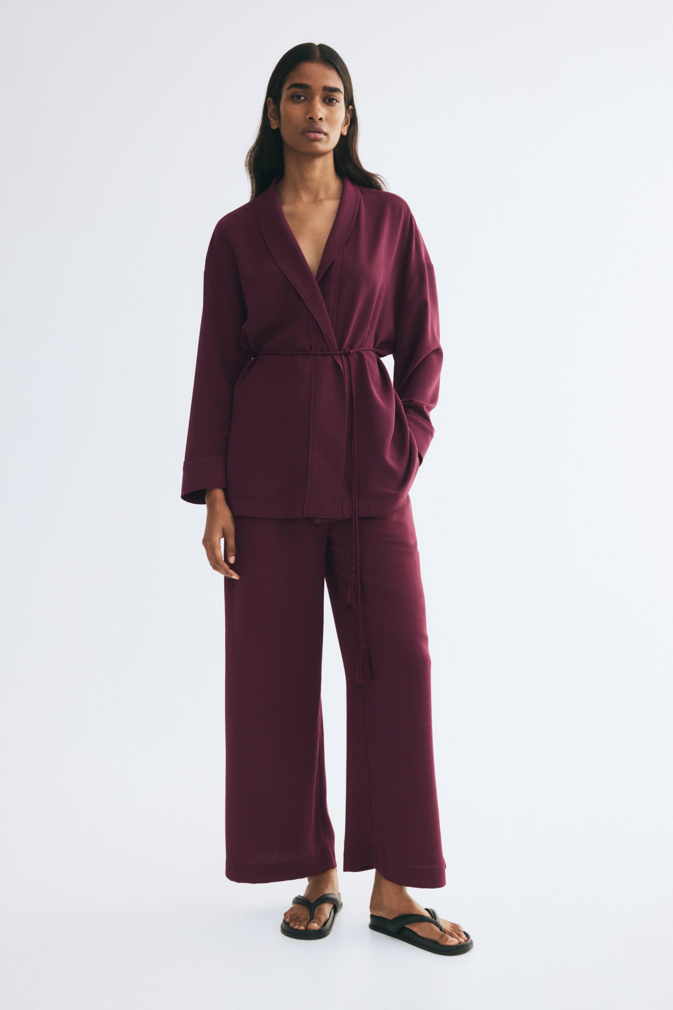View larger image: Relaxed-fit, wide-leg pants in a deep plum woven fabric are being worn by the model, who is standing and facing forward. The pants are styled as a matching set with a long-sleeve belted wrap jacket and black flat slide sandals.