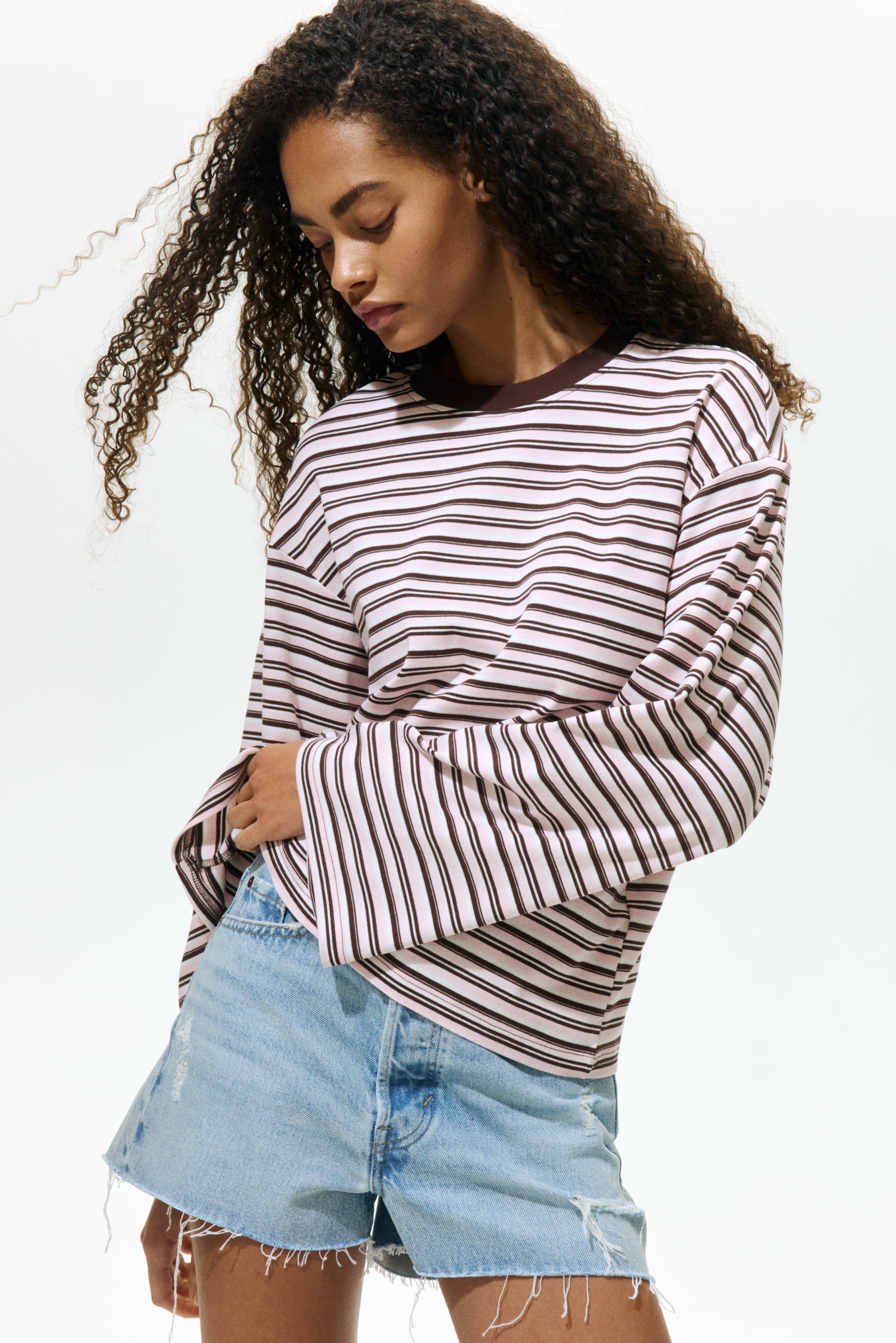Voir une image plus grande: Une chemise à manches longues présente un tissu rose avec de fines rayures horizontales brunes et un col rond côtelé brun. Elle se distingue par des manches larges et amples et une coupe décontractée, partiellement rentrée dans un short en denim bleu clair.
