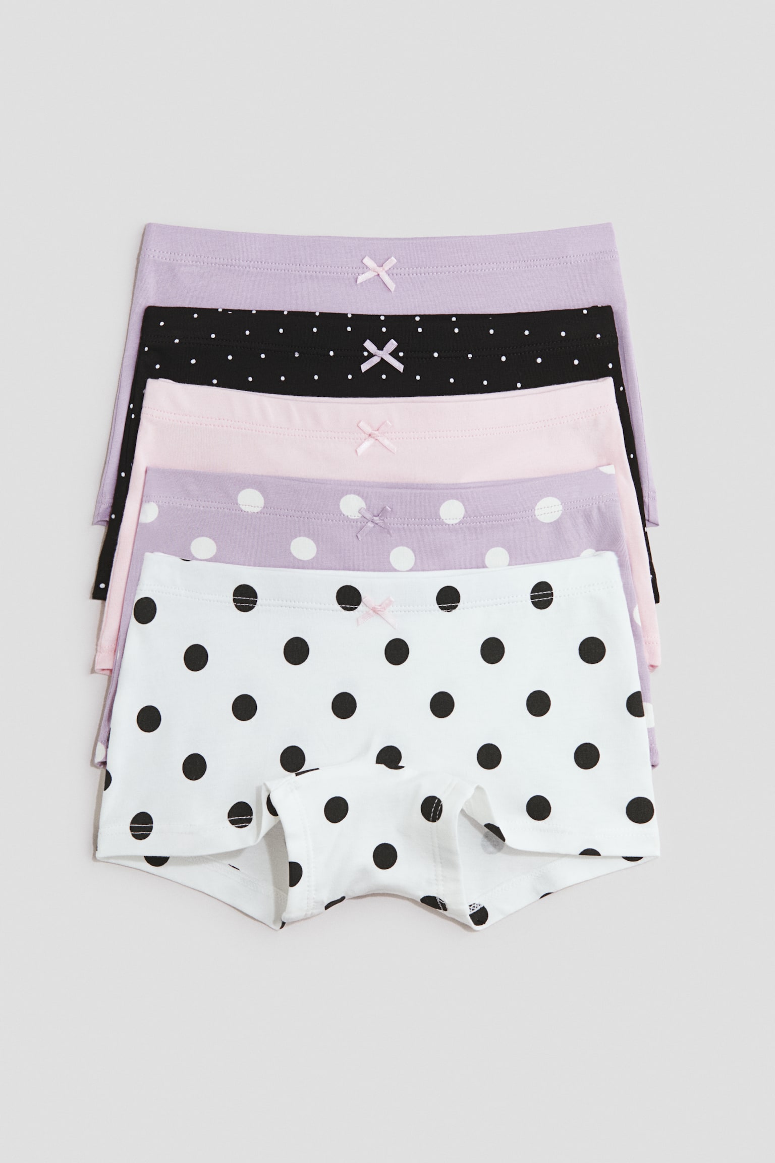 Five cotton jersey boyleg briefs are laid flat in a stacked arrangement, showcasing their designs from the front. Each pair features an elasticated waist with a small decorative bow at the centre, with patterns including solid lilac, black with small white dots, solid light pink, lilac with medium white dots, and white with large black dots.