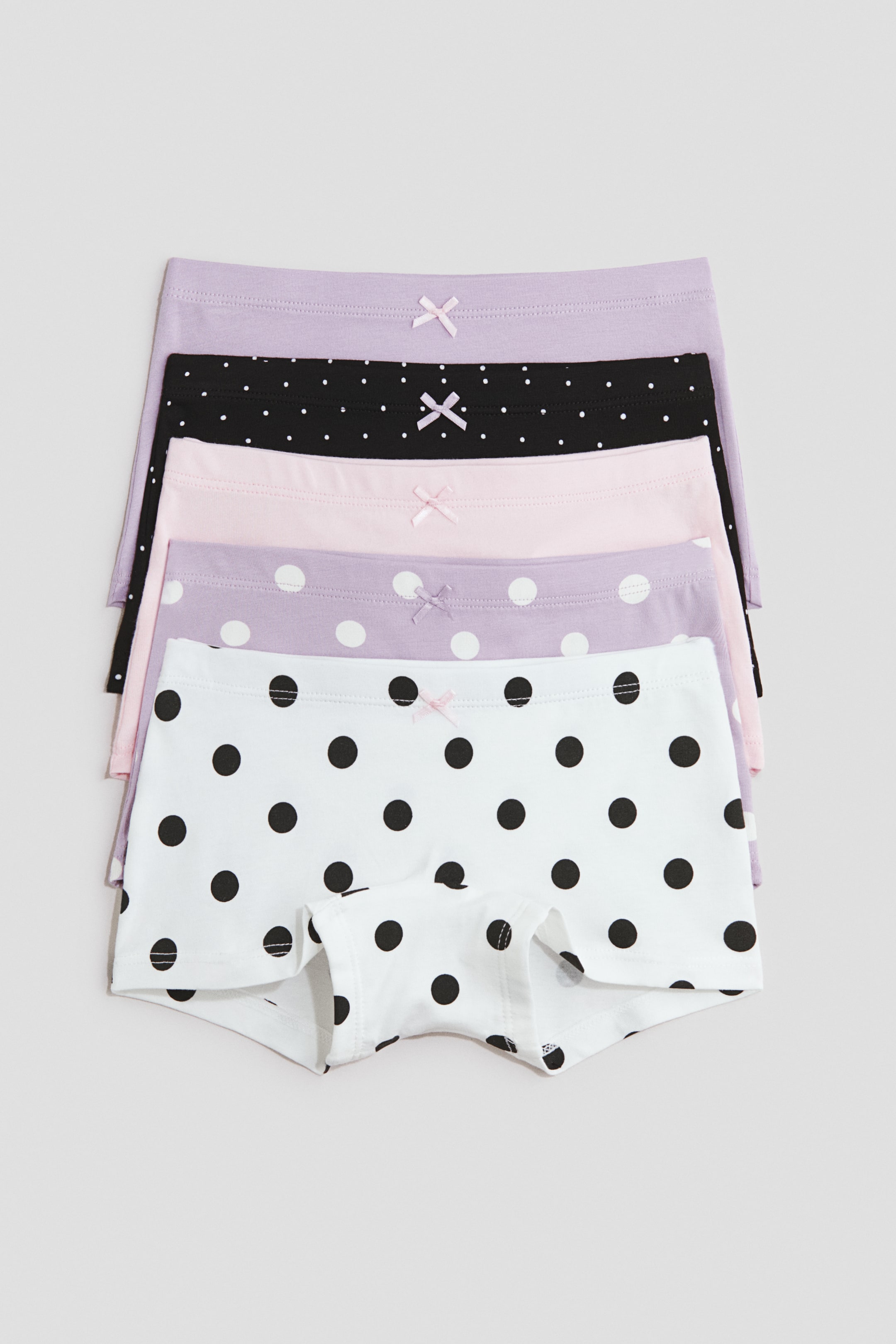 View larger image: Five cotton jersey boyleg briefs are laid flat in a stacked arrangement, showcasing their designs from the front. Each pair features an elasticated waist with a small decorative bow at the centre, with patterns including solid lilac, black with small white dots, solid light pink, lilac with medium white dots, and white with large black dots.