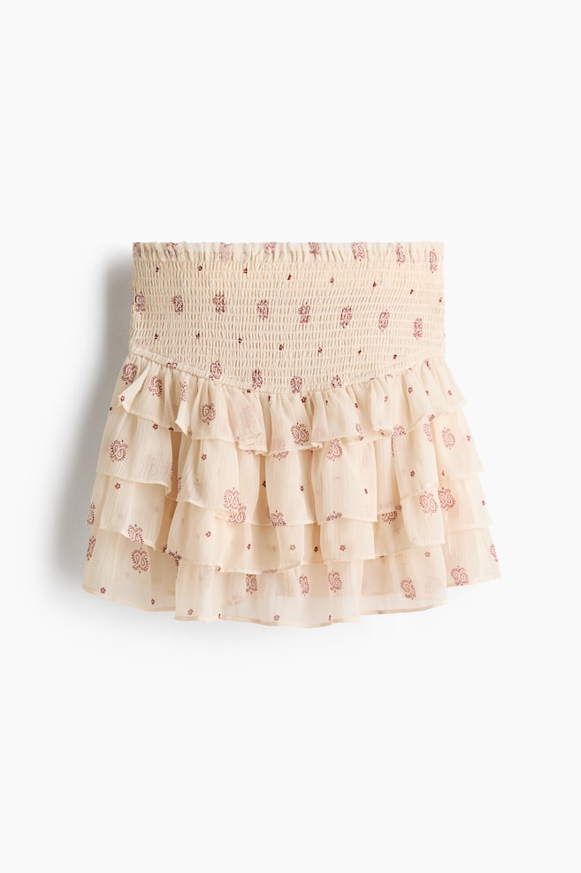 A short skirt is featuring a high, elasticated, and smocked waist, presented from the FRONT. It is made of crinkled chiffon, displaying flounced tiers and a pattern of reddish-brown motifs on a light fabric. - 1