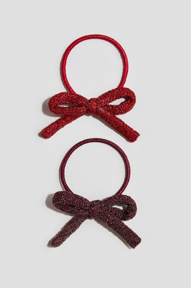 Two elastic hair ties are lying flat, featuring a bright red hair tie on top and a dark burgundy hair tie below. Each hair tie displays a thin, circular elastic band adorned with a matching textured, glittery fabric bow. - 1