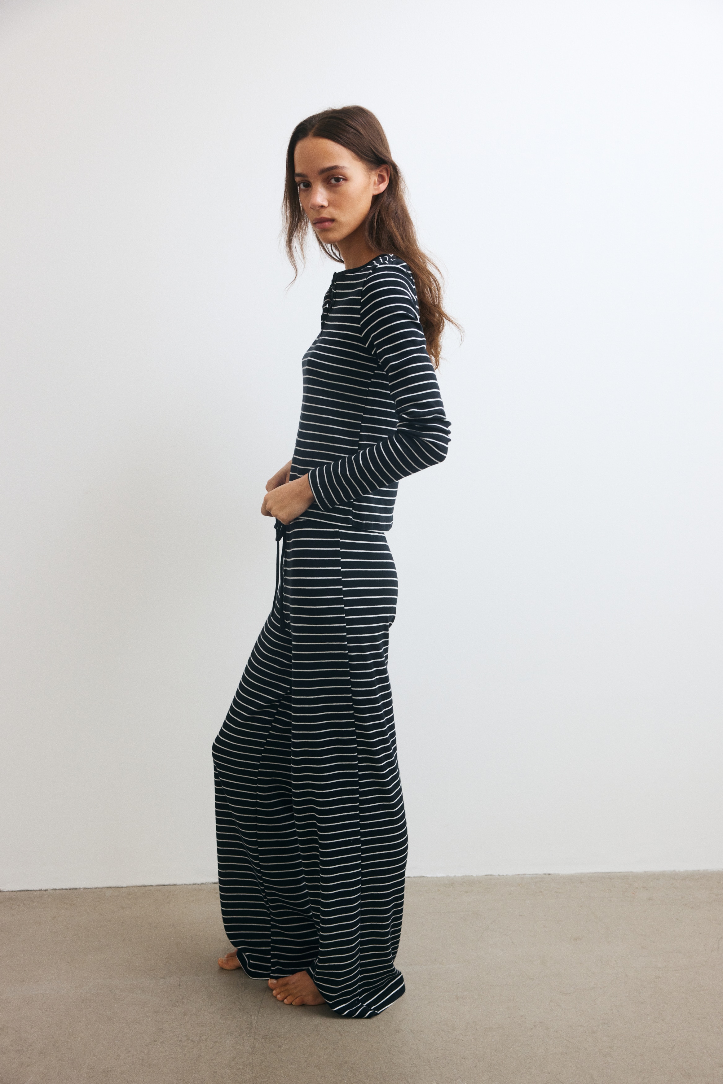 Una vista frontale mostra un pigiama a righe orizzontali bianche e nere in maglia pointelle dalla texture lavorata. Il completo include un top a maniche lunghe con scollo rotondo e apertura con bottoni, abbinato a un pantalone dritto coordinato.