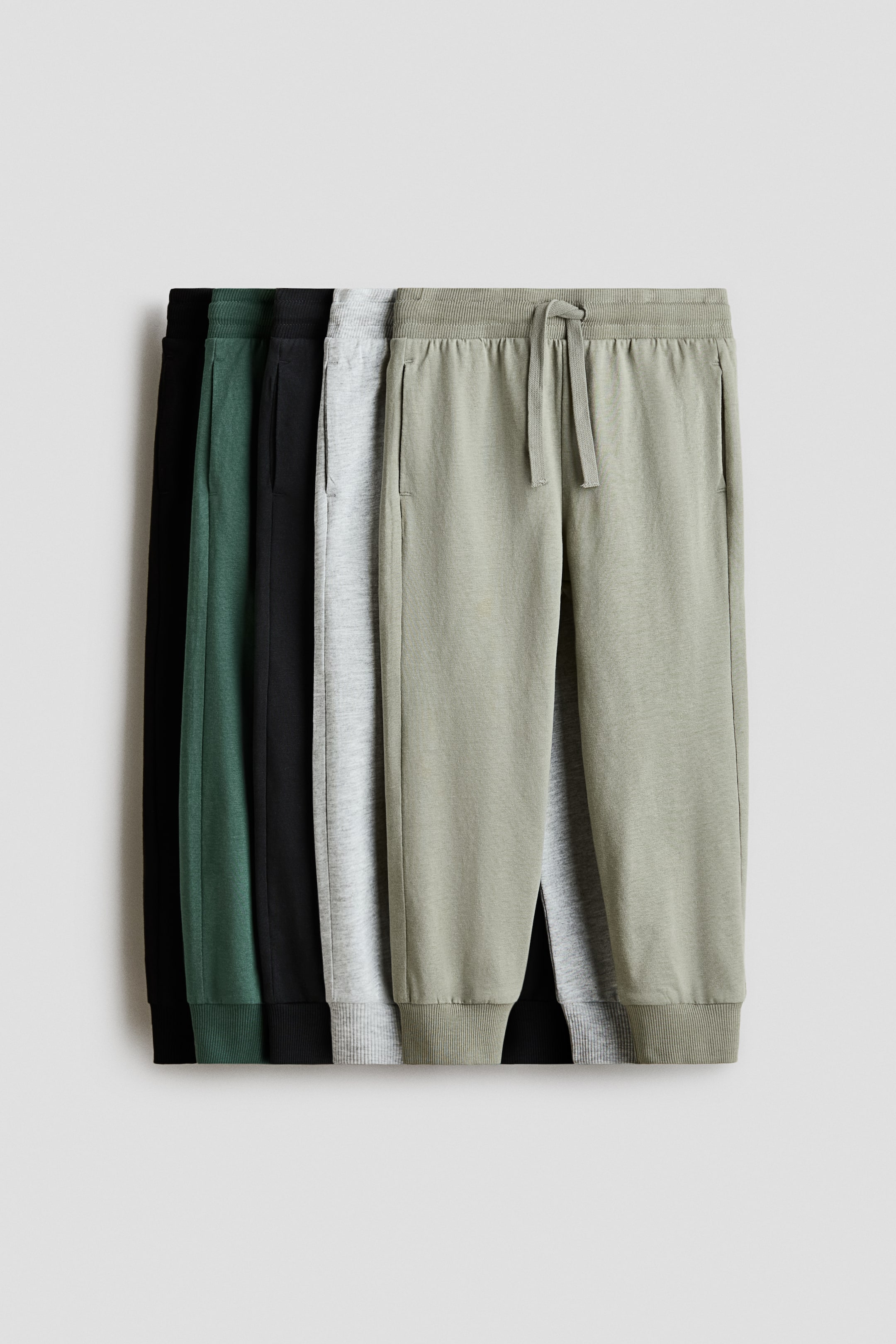 Ver imagem maior: Joggers exibem a FRENTE, com cintura elástica com cordão, bolsos laterais discretos e bainhas com punhos canelados. Vários pares em verde-claro, cinzento-claro, verde-escuro e preto estão parcialmente visíveis, empilhados uns sobre os outros.