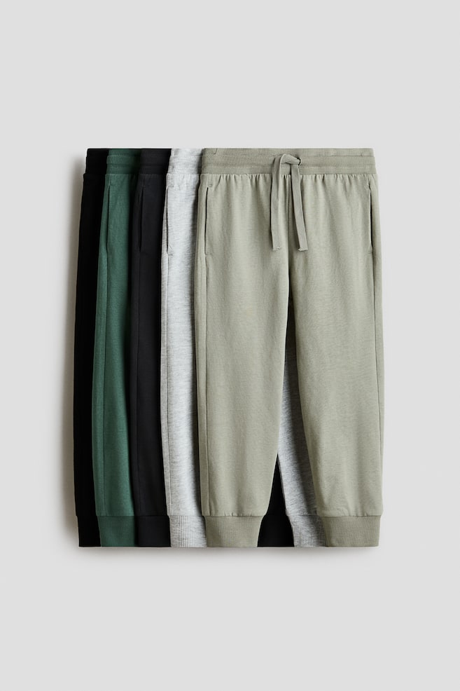 Joggers are displaying their FRONT, featuring an elasticated, drawstring waist, discreet side pockets, and ribbed cuffs at the hems. Multiple pairs in light green, light gray, dark green, and black are partially visible, stacked behind each other. - 1