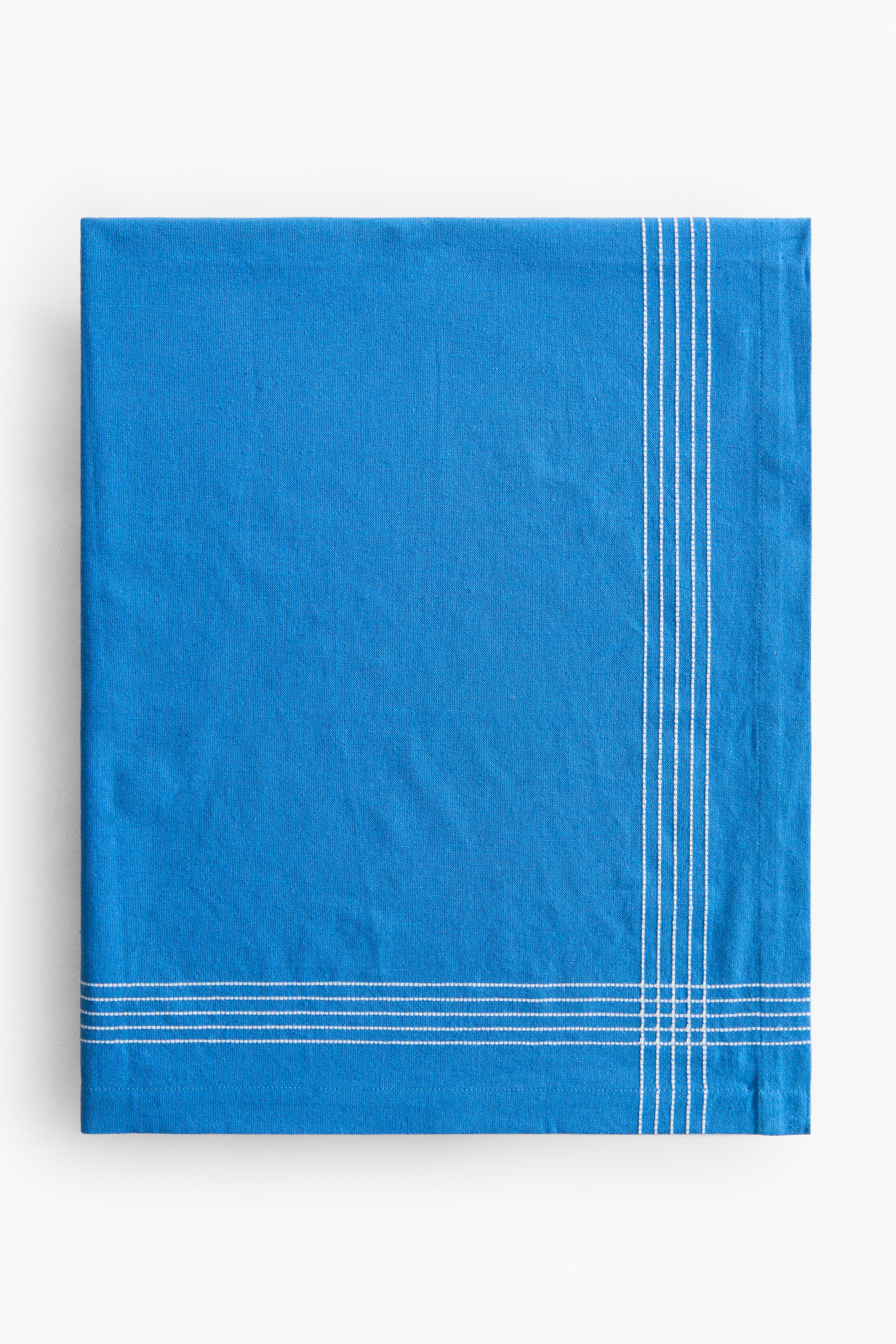 Ver imagen más grande: Un paño cuadrado azul está extendido, mostrando una textura tejida sutil y ligeras arrugas. Presenta tres finas rayas blancas verticales a lo largo del borde derecho y tres finas rayas blancas horizontales a lo largo del borde inferior, que se intersectan para formar una cuadrícula en la esquina inferior derecha.