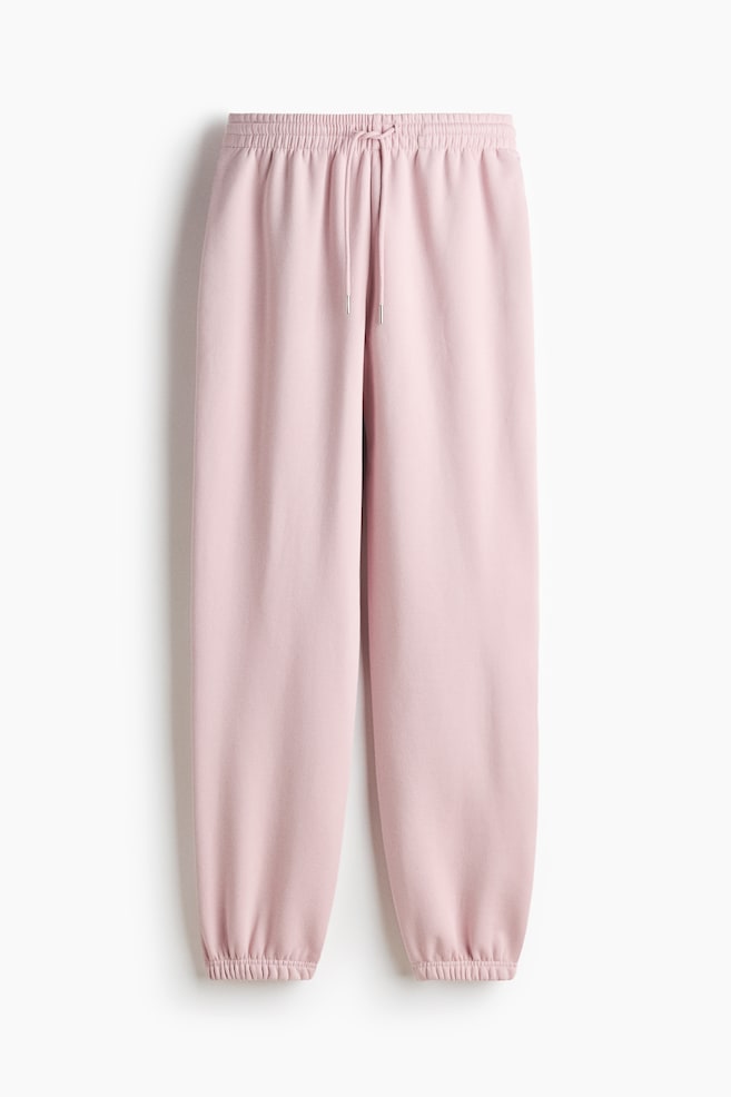 Los pantalones de chándal rosados se muestran en vista frontal, hechos de una suave tela de sudadera, con una cintura alta elástica y un cordón central con puntas plateadas, y dobladillos elásticos en los tobillos. - 1