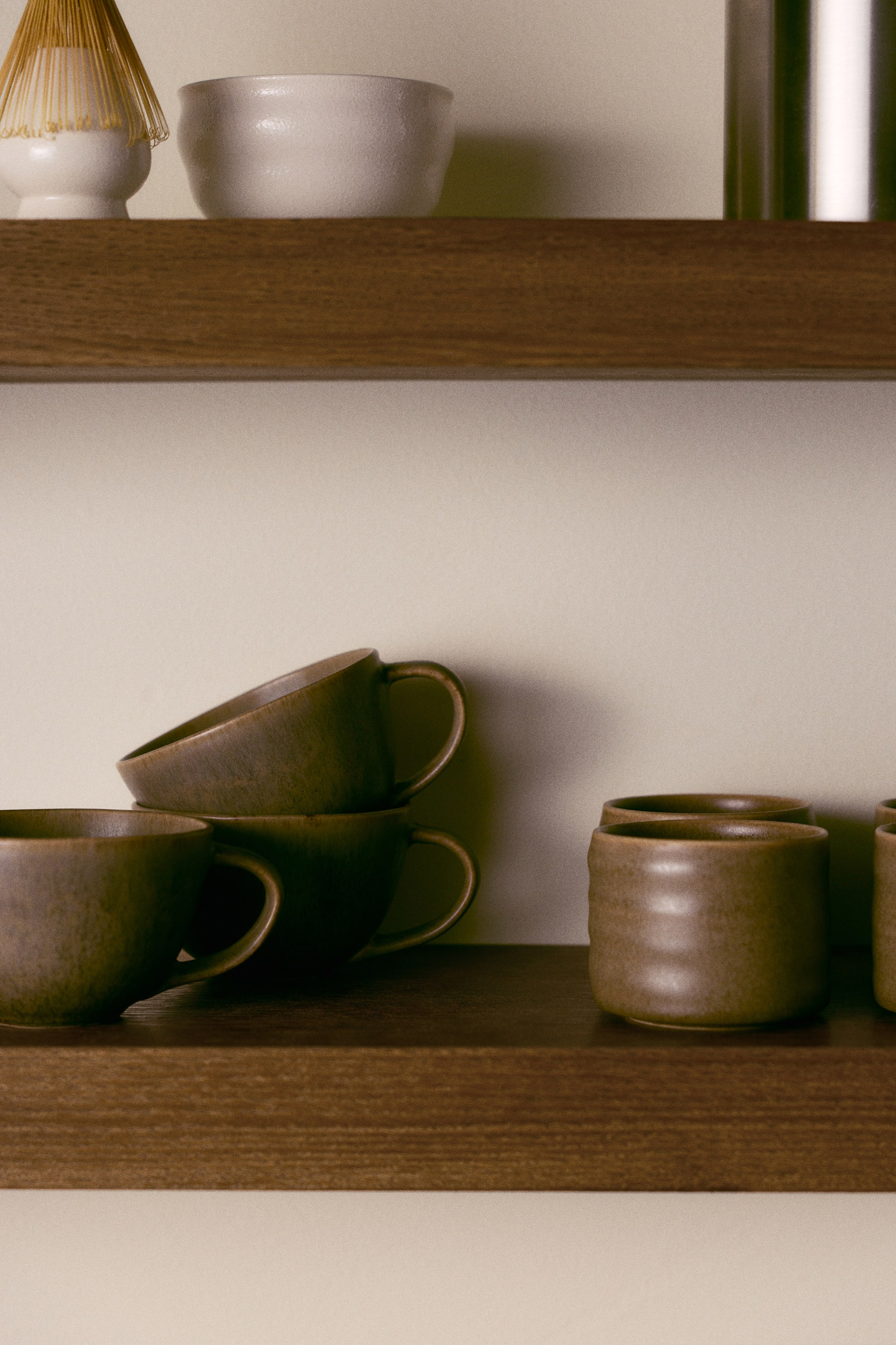 View larger image: Stoneware tea mugs finished with an olive-green reactive glaze rest on a wooden shelf, with one mug placed in the foreground and two others stacked behind it. The mugs feature a broad, rounded silhouette, curved loop handles, and a unique, subtly textured finish.