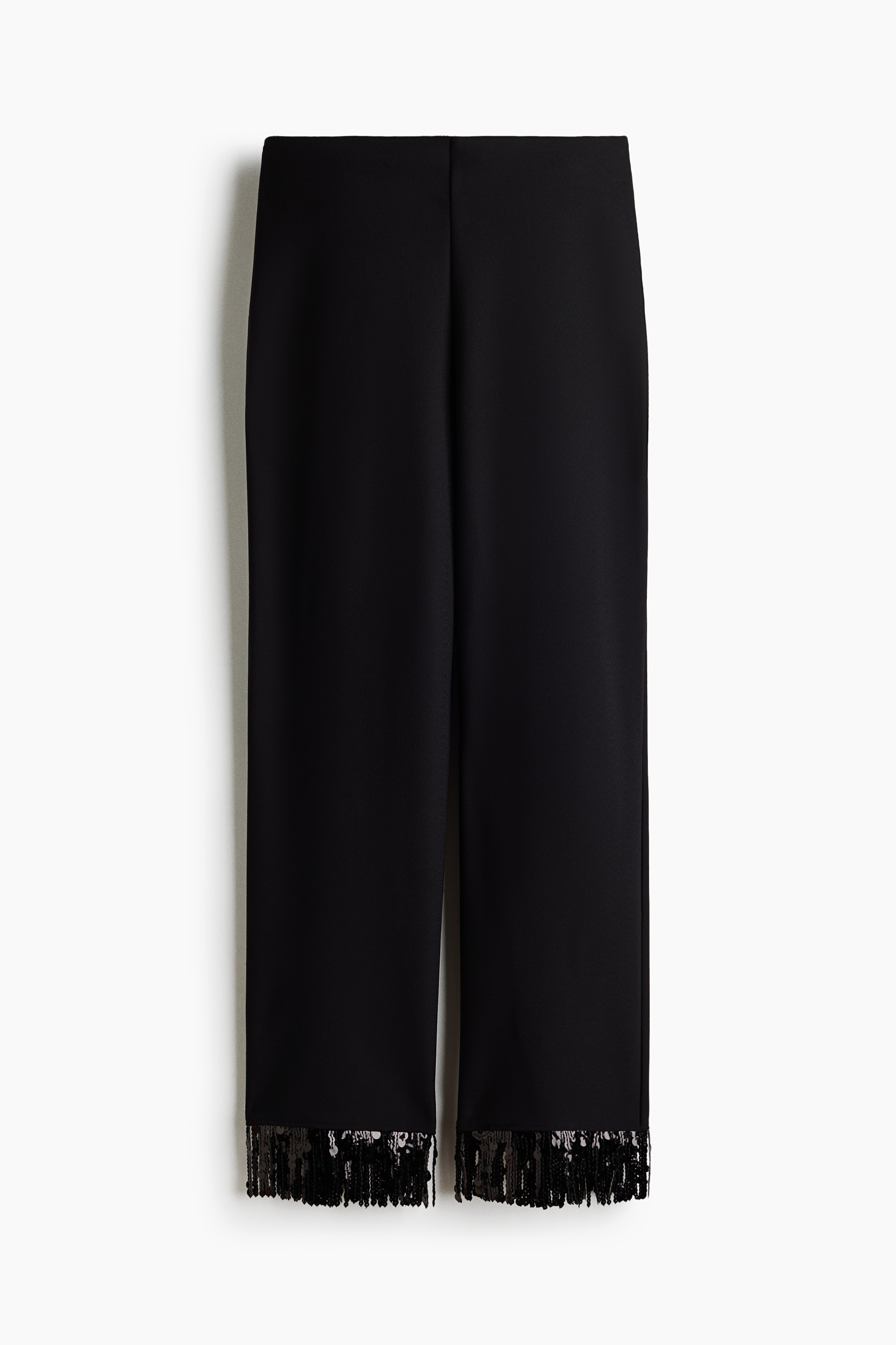 A black sleeveless top featuring a sequin-embellished fringe hem is being worn by the model with matching black pants that also have a fringed hem. The coordinated ensemble is styled with black strappy heeled sandals.