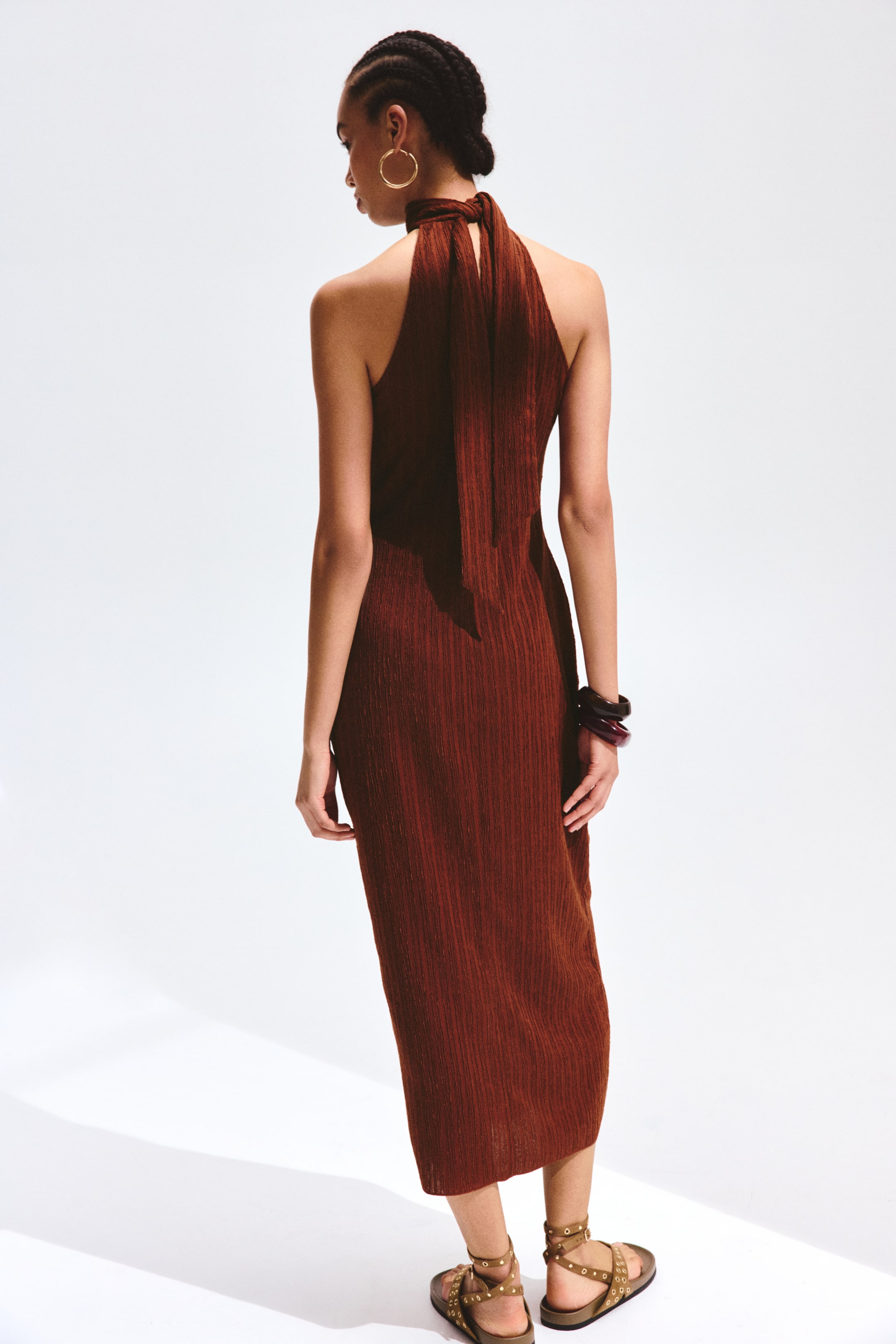 View larger image: A rust-coloured midi dress featuring a halter-neck design with ties at the back of the neck. The dress showcases a finely pleated vertical texture throughout and closely fits the body, as viewed from the back.
