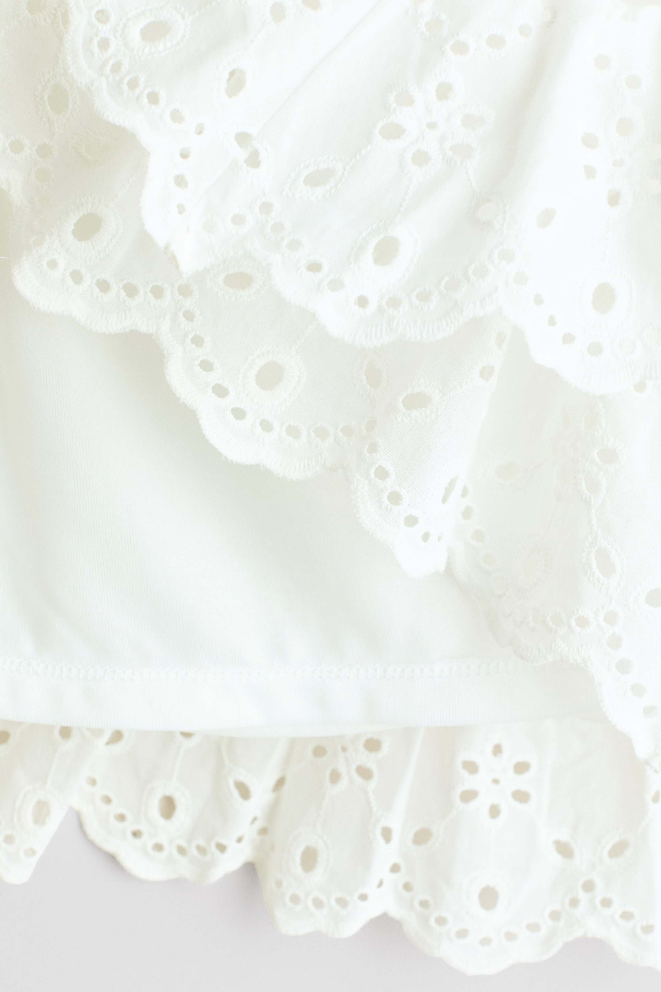 Ver imagen más grande: Una falda blanca presenta un tejido de algodón aireado con broderie anglaise en capas, cada nivel mostrando bordes festoneados y patrones de ojales intrincados. La falda también revela un forro liso y sencillo debajo del broderie anglaise.