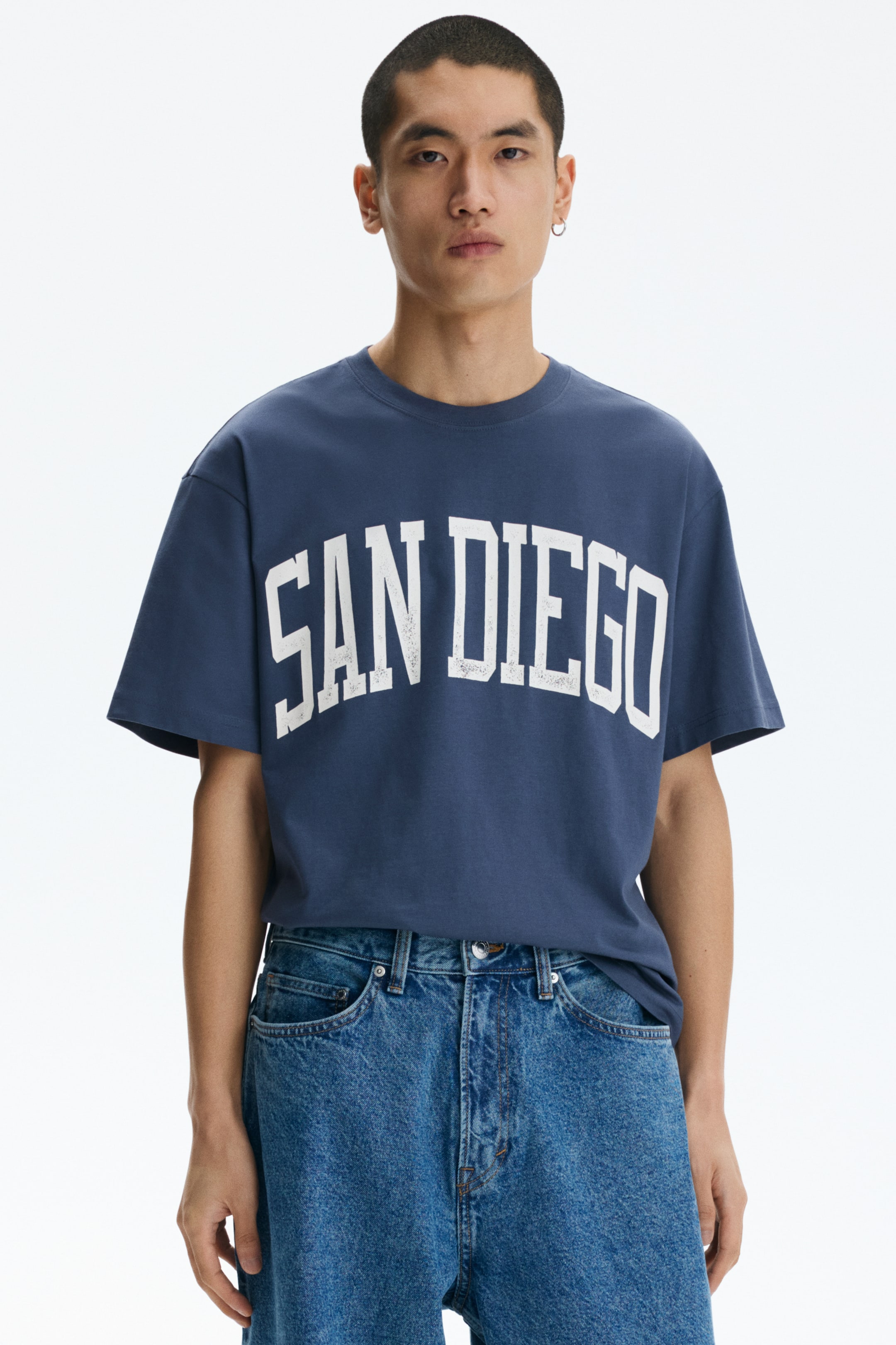 Voir une image plus grande: Un t-shirt à col rond bleu marine arbore "SAN DIEGO" en lettres blanches usées et arquées sur la poitrine. Le t-shirt ample est porté avec l'avant rentré dans un jean bleu clair.