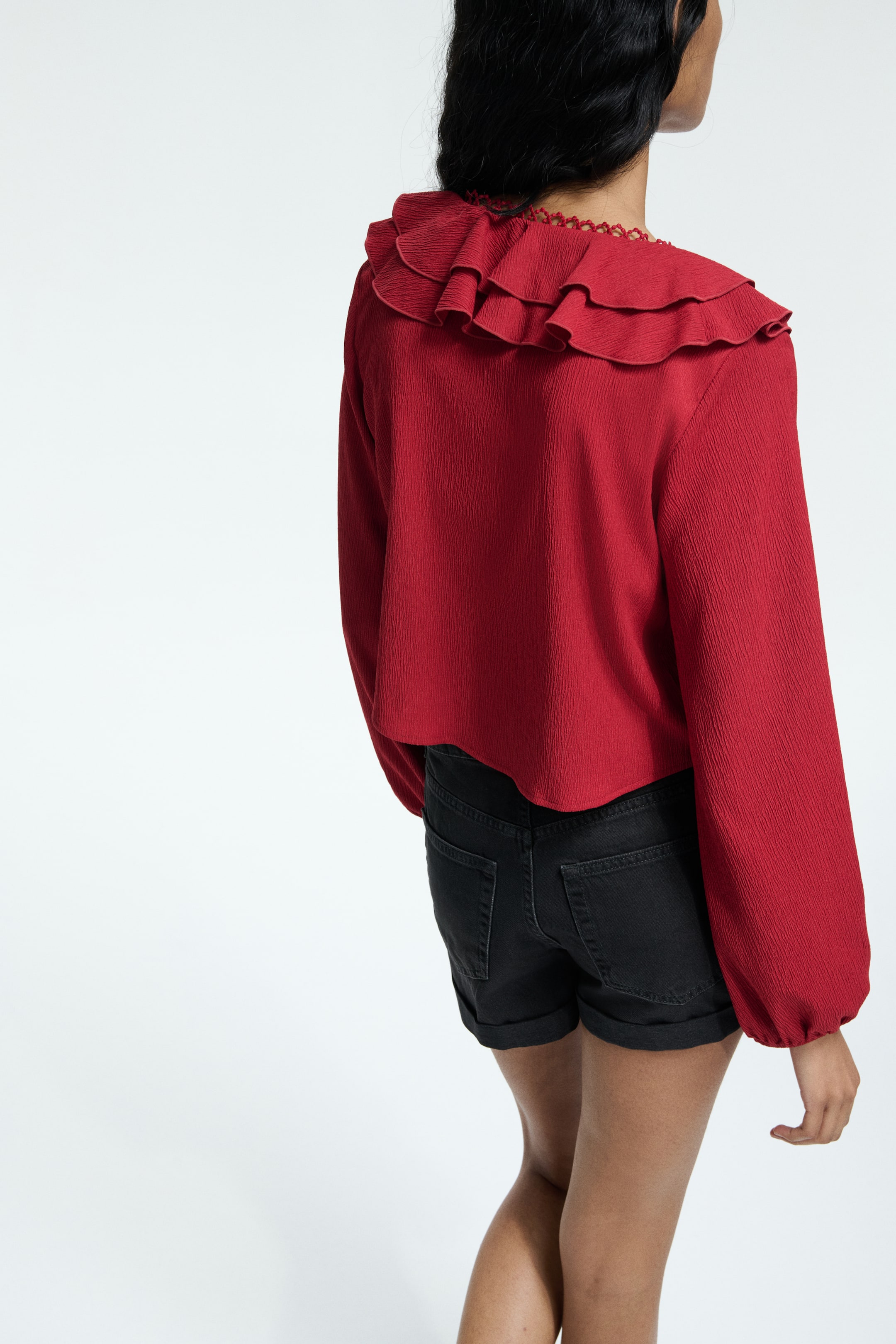 View larger image: A red blouse is being viewed from the back, angled slightly to the right, featuring a crinkled texture. It is showcasing a large, layered ruffle collar around the neckline and shoulders, a delicate trim at the neck, and long, full sleeves, with its hem ending just above the waist.