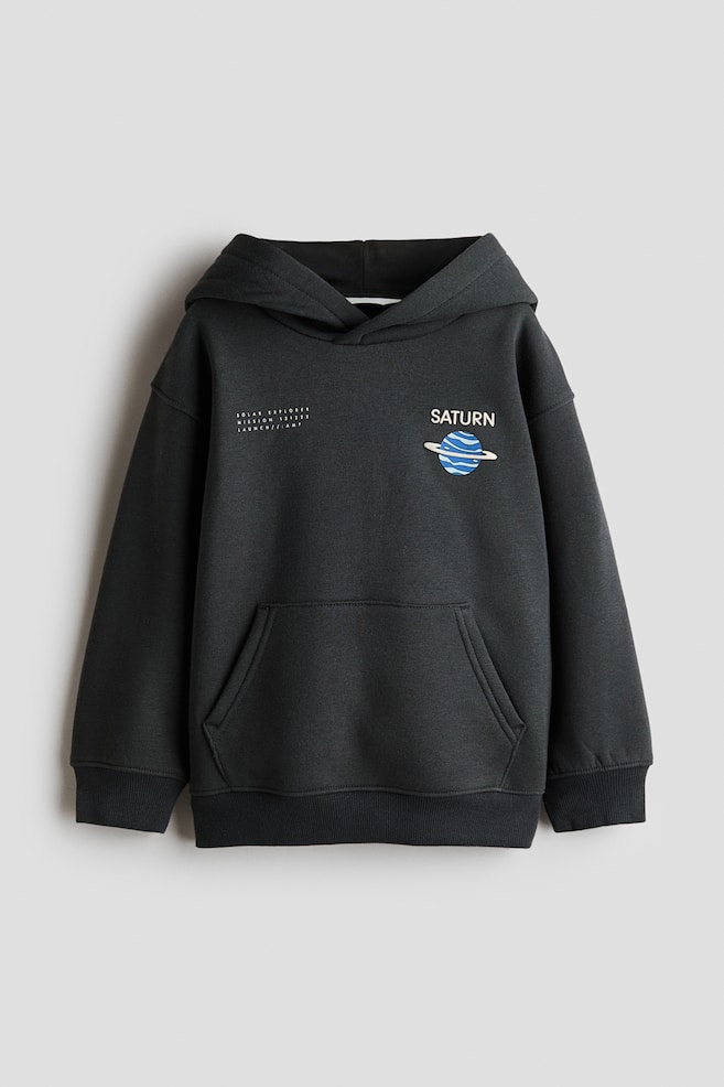 This front-facing dark grey loose-fit hoodie is featuring a jersey-lined hood with a wrap front, dropped shoulders, and a front kangaroo pocket. The chest is displaying a small graphic of the planet Saturn on the left and a small block of white text on the right, while wide ribbed trims are finishing the cuffs and hem. - 1