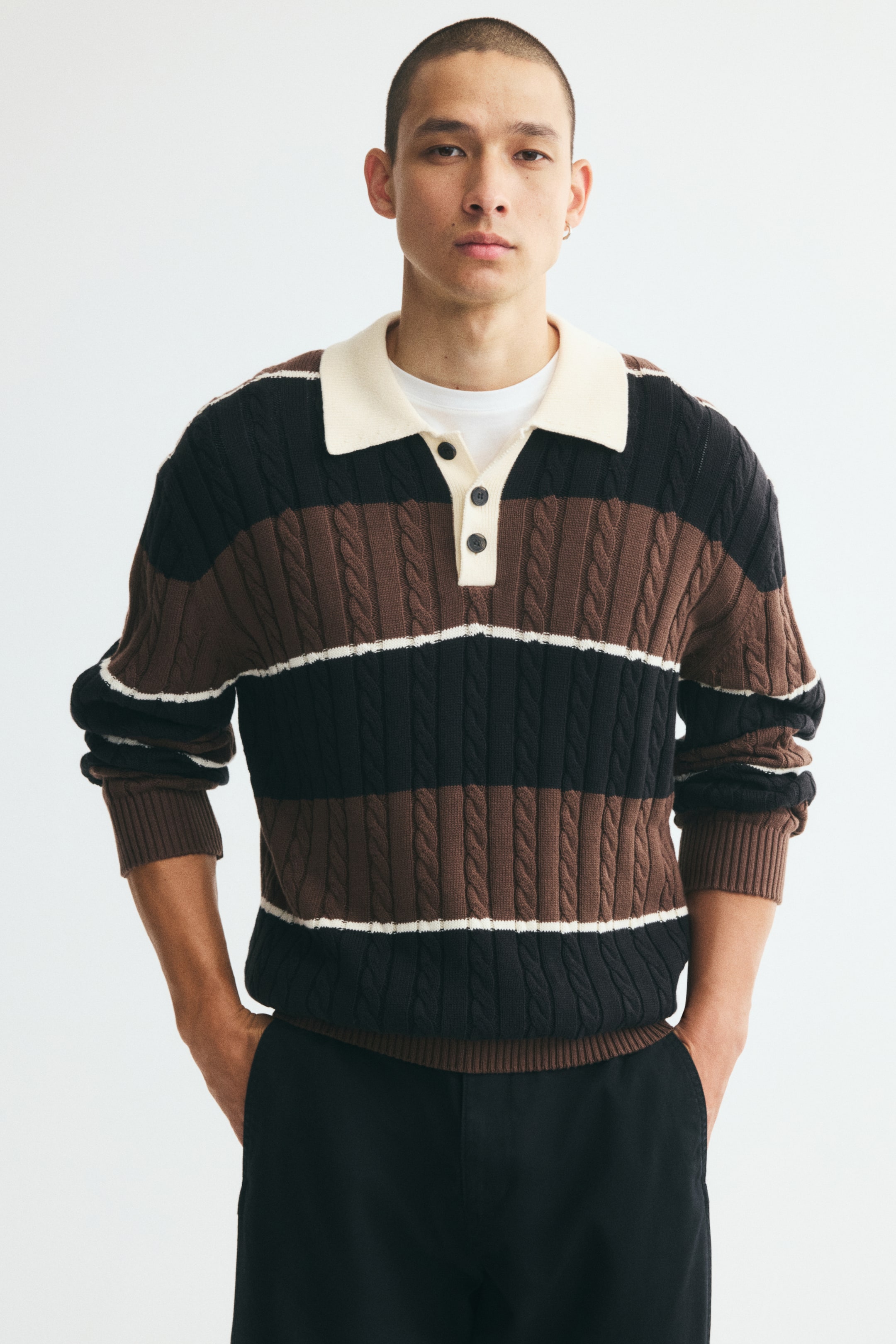 Voir une image plus grande: Un polo à manches longues à coupe décontractée, au design tricot torsadé avec des rayures horizontales brunes, noires et blanches, est porté par le mannequin. Le polo présente un col contrastant de couleur crème, une patte de boutonnage courte ainsi que des poignets et un ourlet côtelés, superposé à un t-shirt blanc et agencé avec un pantalon noir.