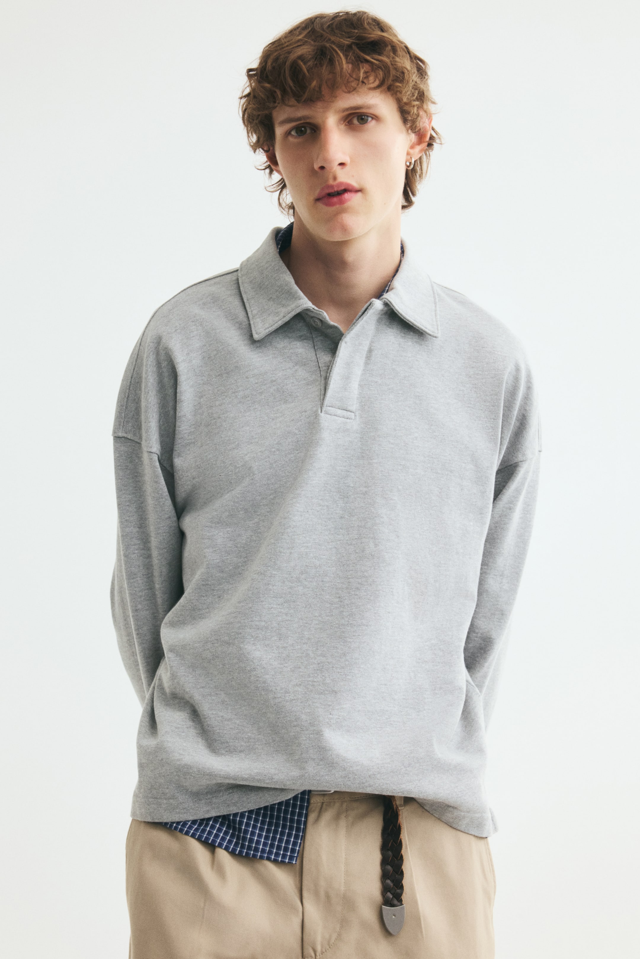 Visa större bild: En ljusgrå långärmad pikétröja bärs av modellen och har en oversized passform, sänkta axlar, klassisk krage och dold knappslå. Pikétröjan är lager på lager över en blå rutmönstrad skjorta och stylad med beige byxor.
